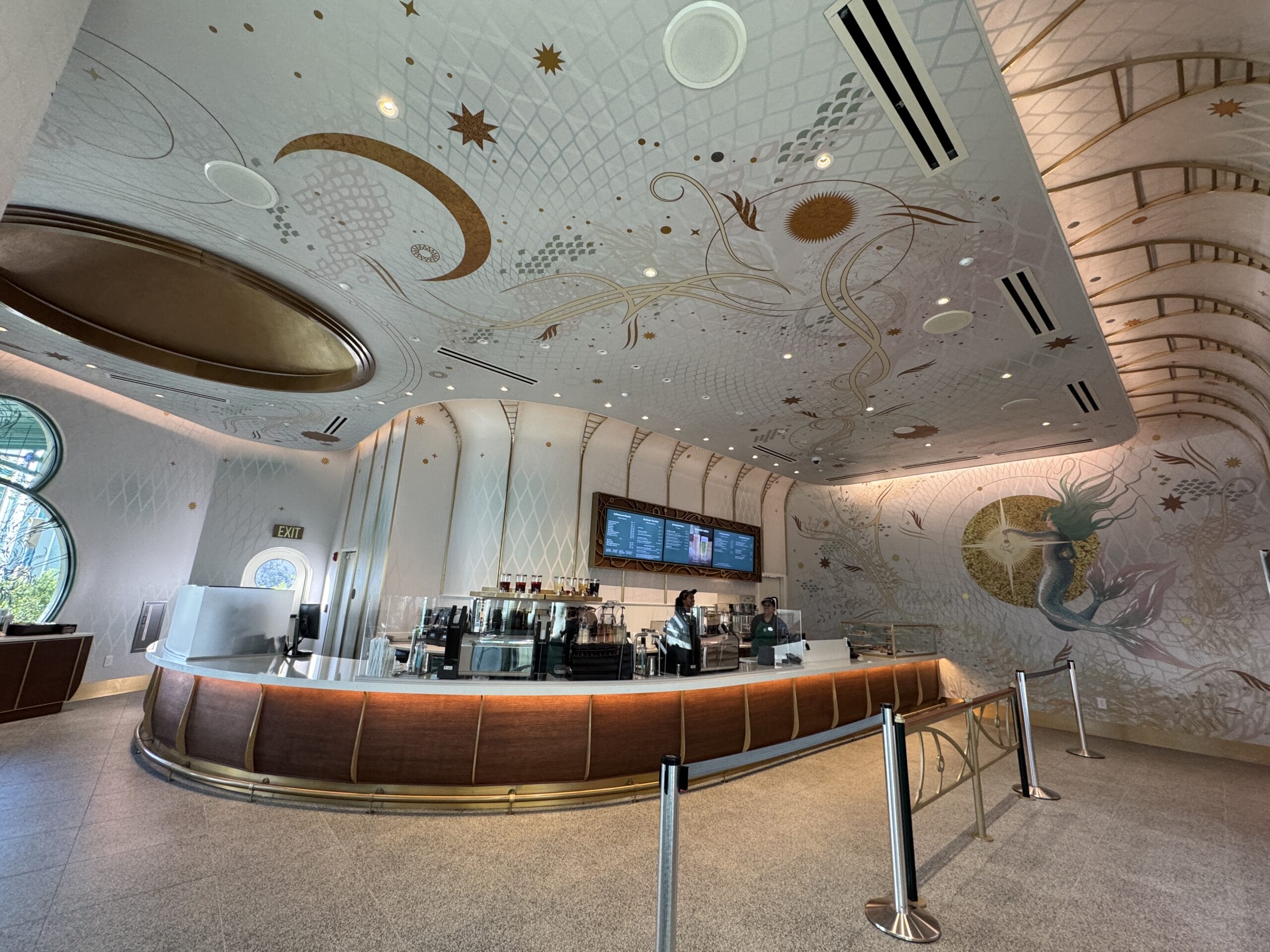 A whimsical café interior reminiscent of Fantasyland in Disneyland, with a mermaid mural, ornate ceiling, and two staff behind counter.