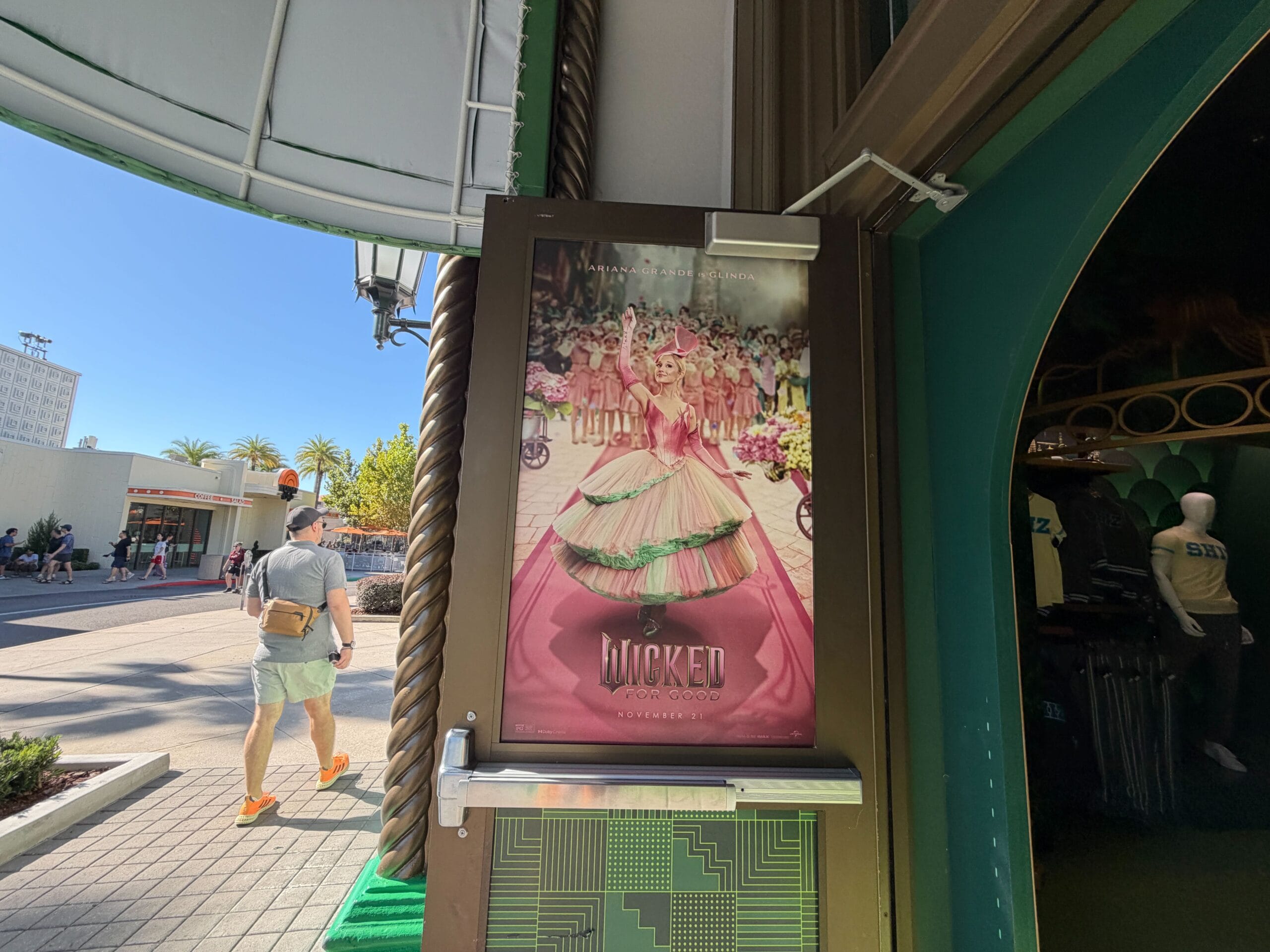 A Wicked poster with a woman in a pink dress shows off vibrant costumes outside a shop in bright sunshine, similar to Main Street’s displays.