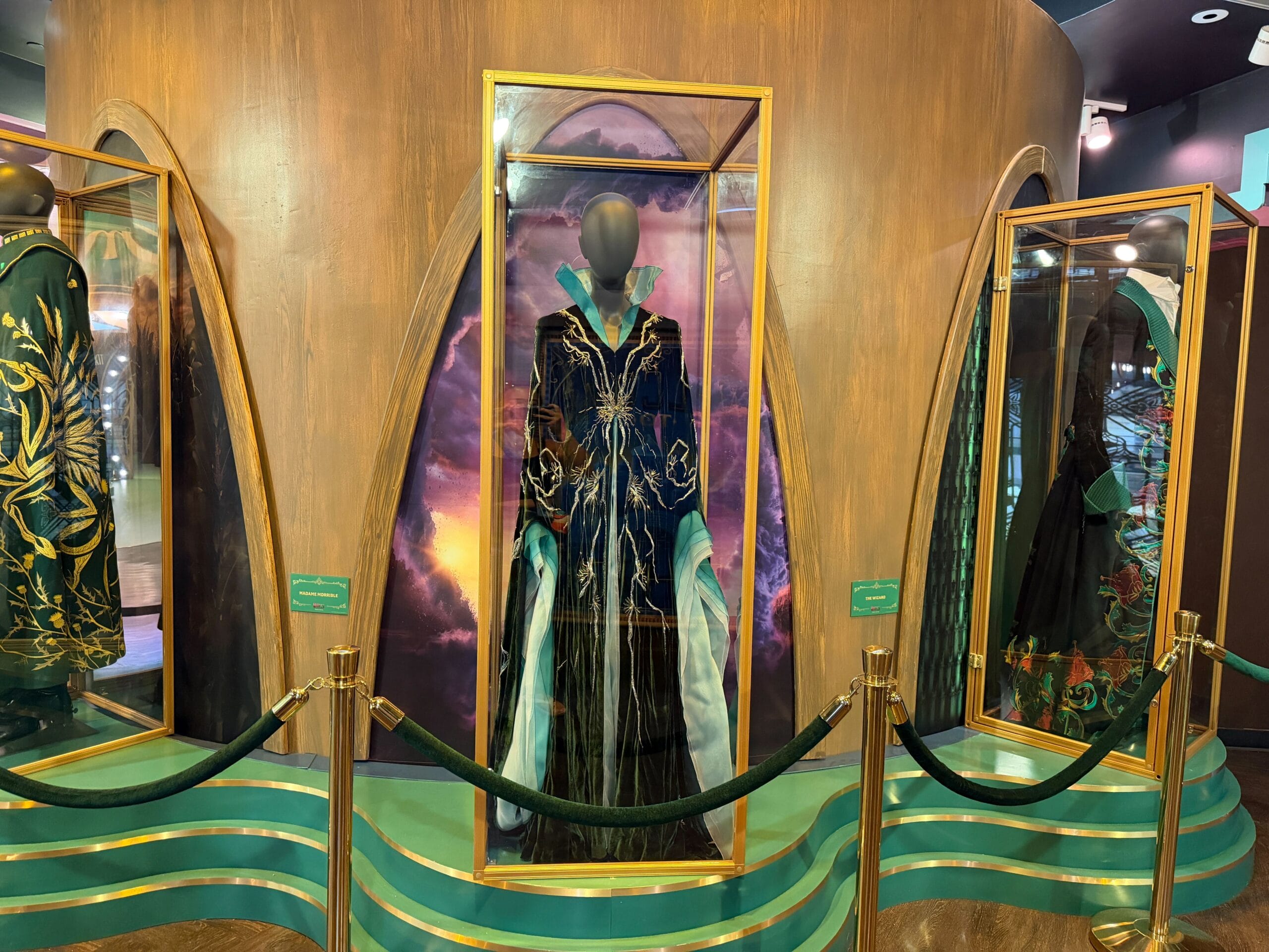A black, gold, and green ornate Wicked robe is showcased with a magical backdrop, evoking the grandeur of Disney park castles.