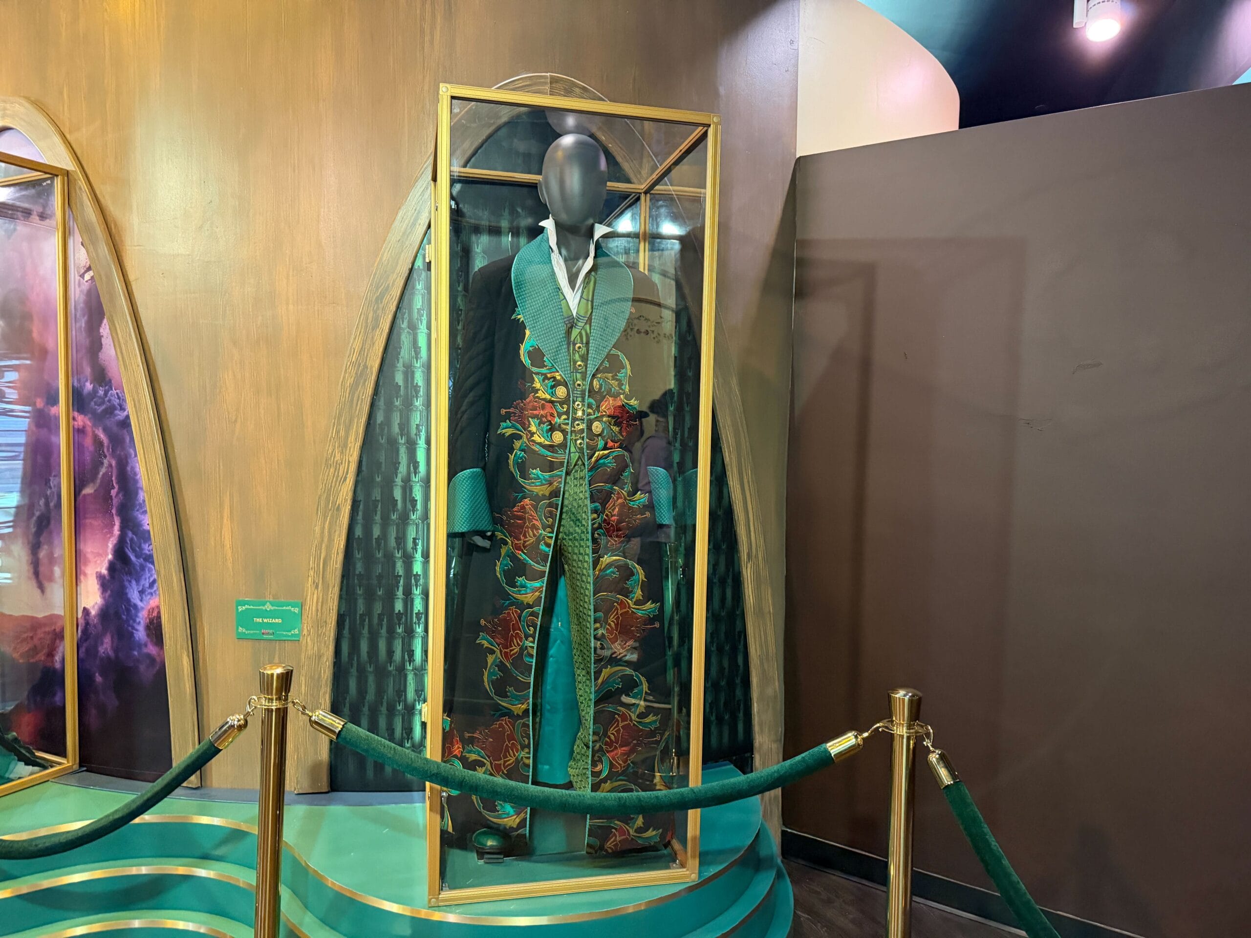 A mannequin in a glass case showcases an ornate green robe from the Wicked Experience, reminiscent of detailed costumes at Disney Parks.
