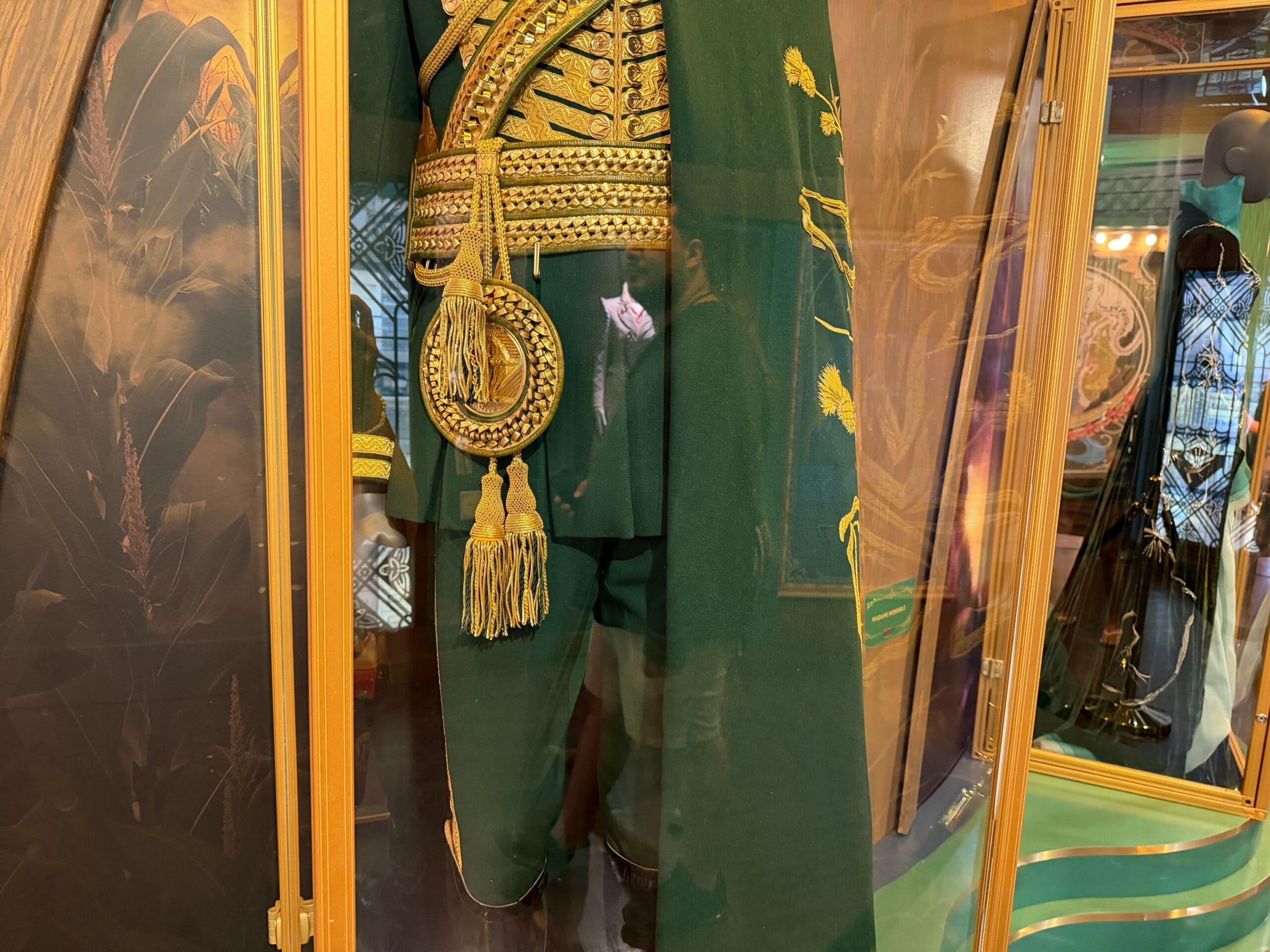 A green military-style costume with gold embroidery, similar to Wicked, displayed in a glass case at a Disney Park exhibit.
