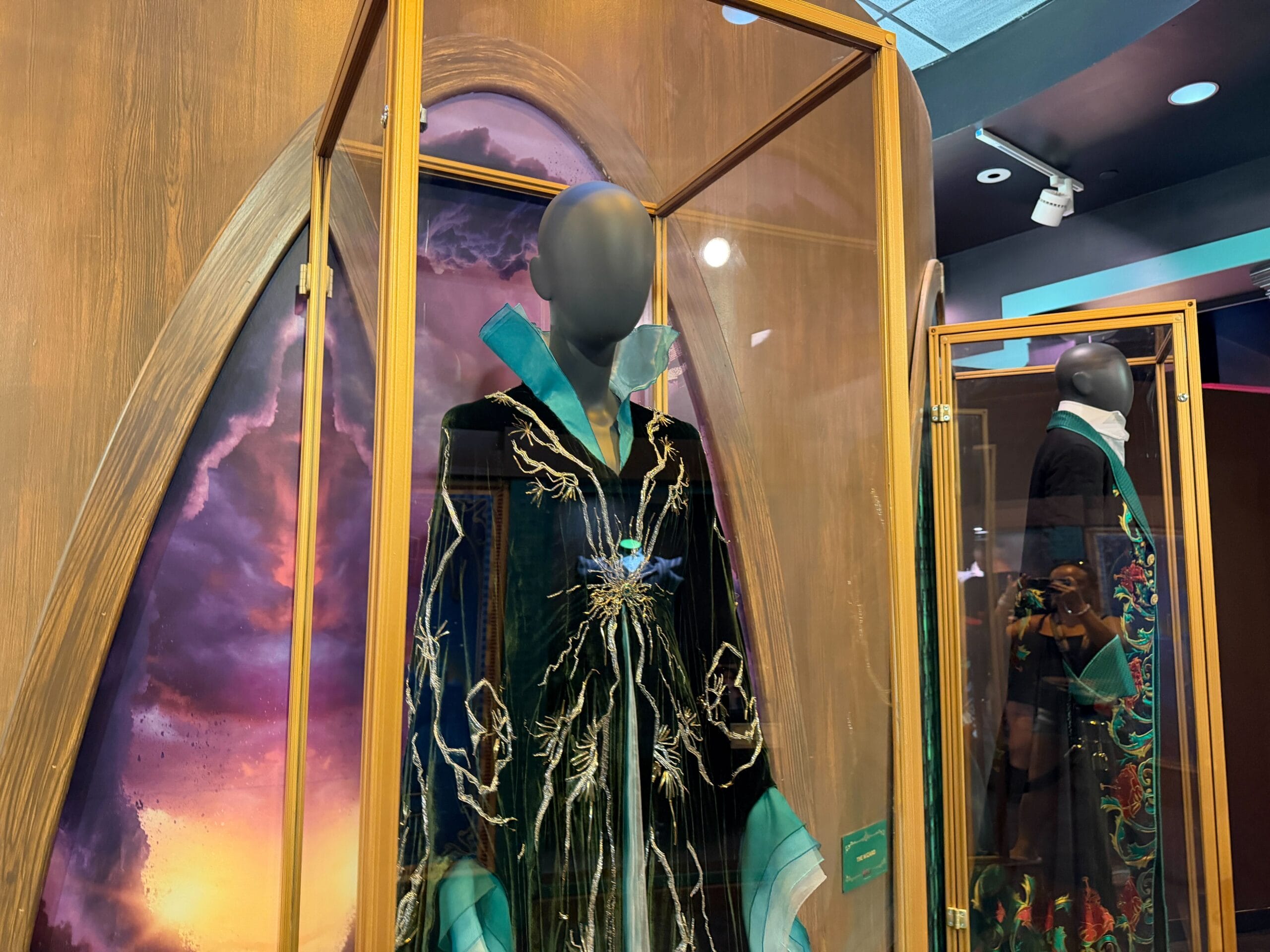 A mannequin in a glass case showcases an ornate black robe with blue and gold embroidery, reminiscent of elaborate Disney park costumes.
