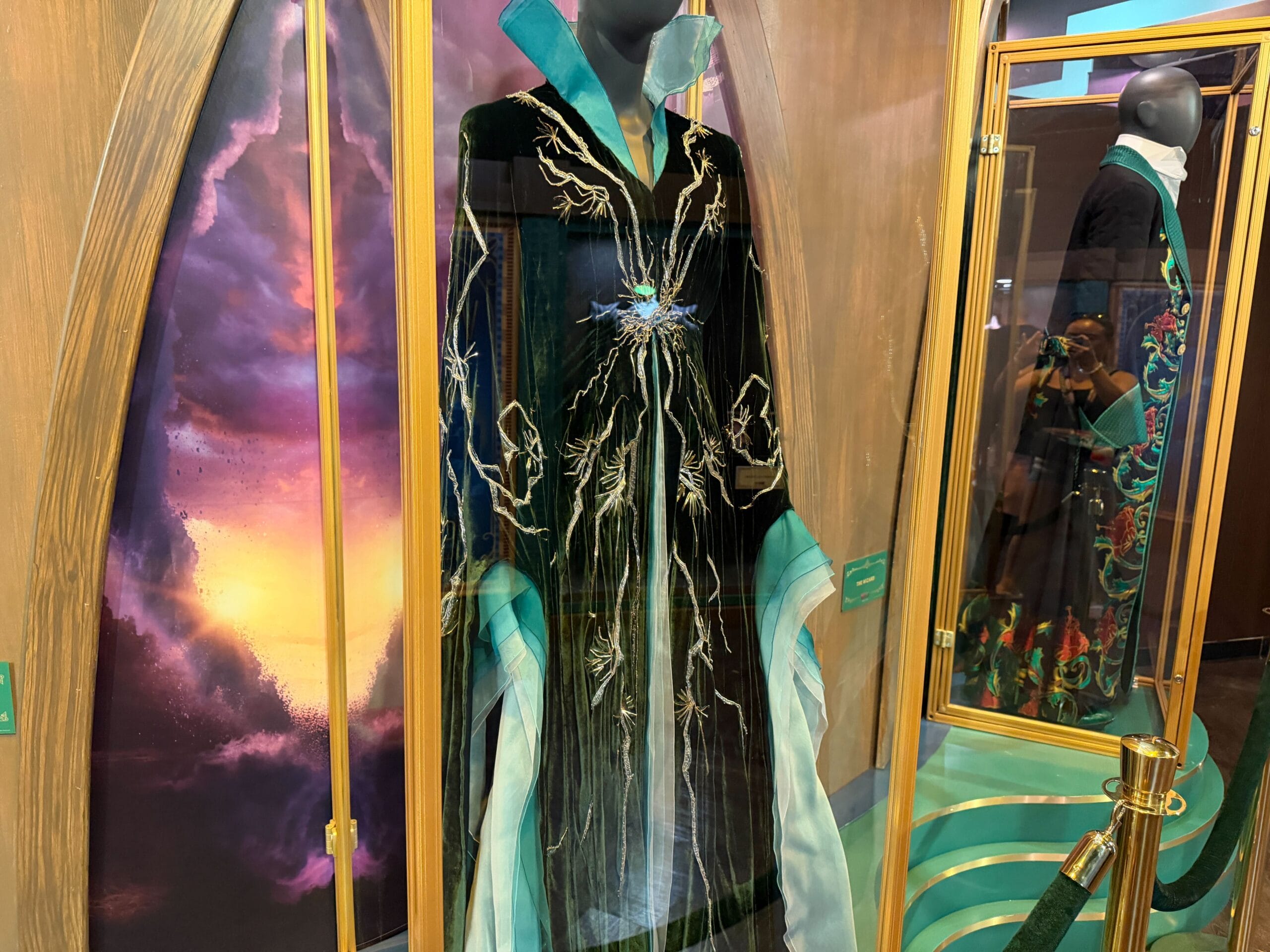 A mannequin shows a black embroidered gown with turquoise details, displayed by Wicked costumes near a backdrop reminiscent of Disney parks.