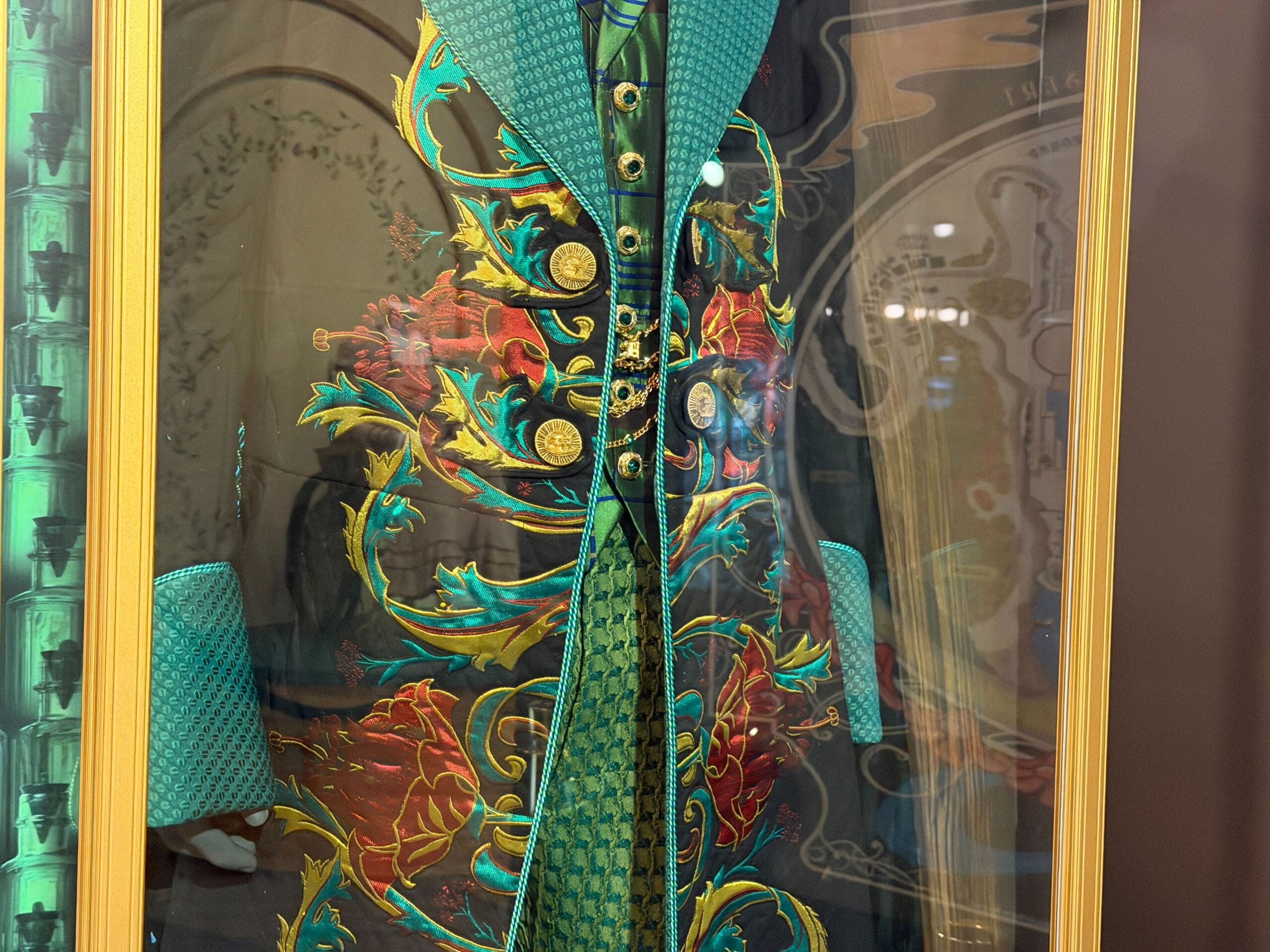 A vibrant teal and green coat with gold buttons, reminiscent of elaborate Disney parade costumes, displayed in a glass case.