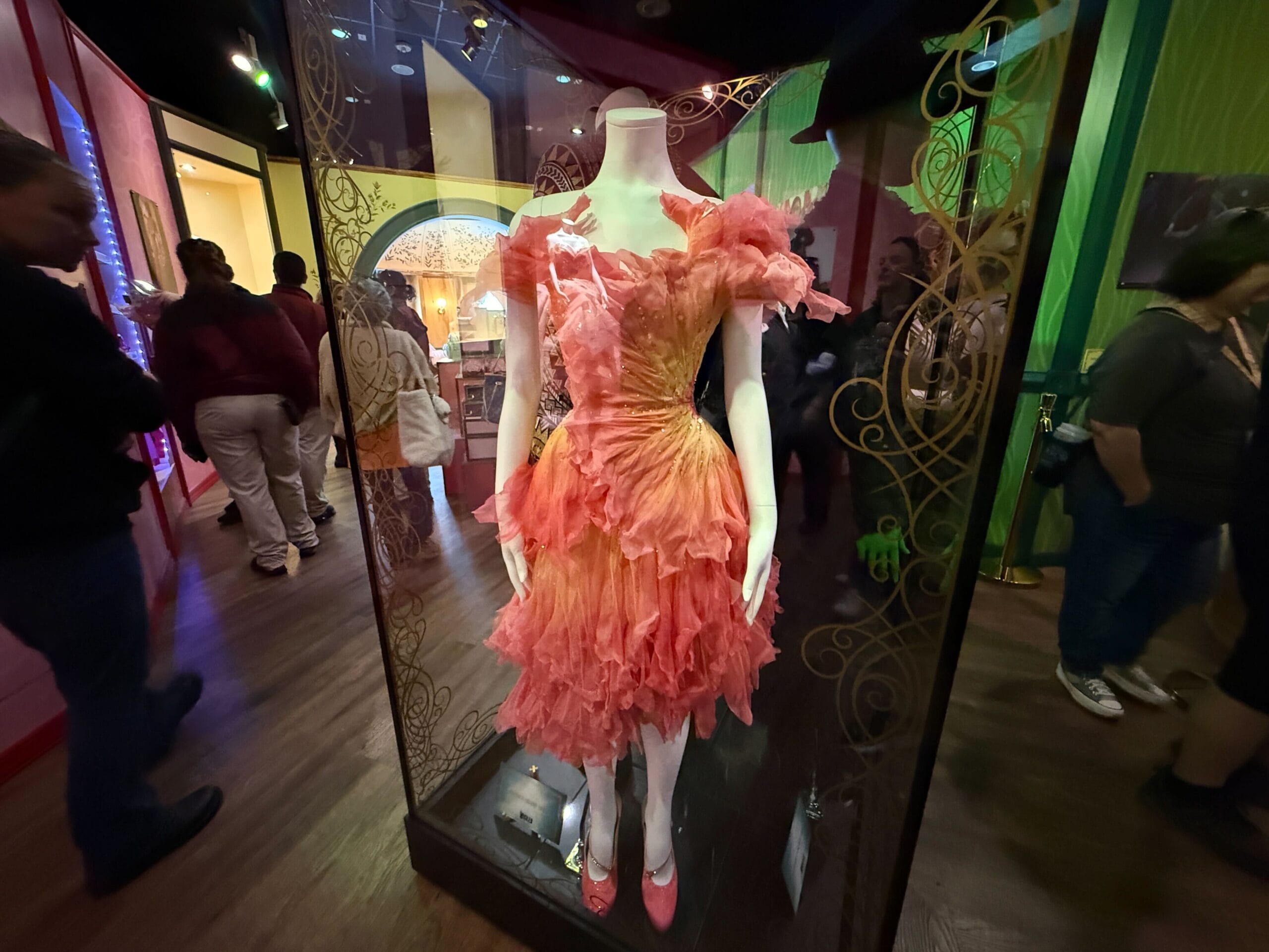 A mannequin displays a ruffled orange and pink dress in a glass case at a Disney Parks exhibit, with visitors nearby.