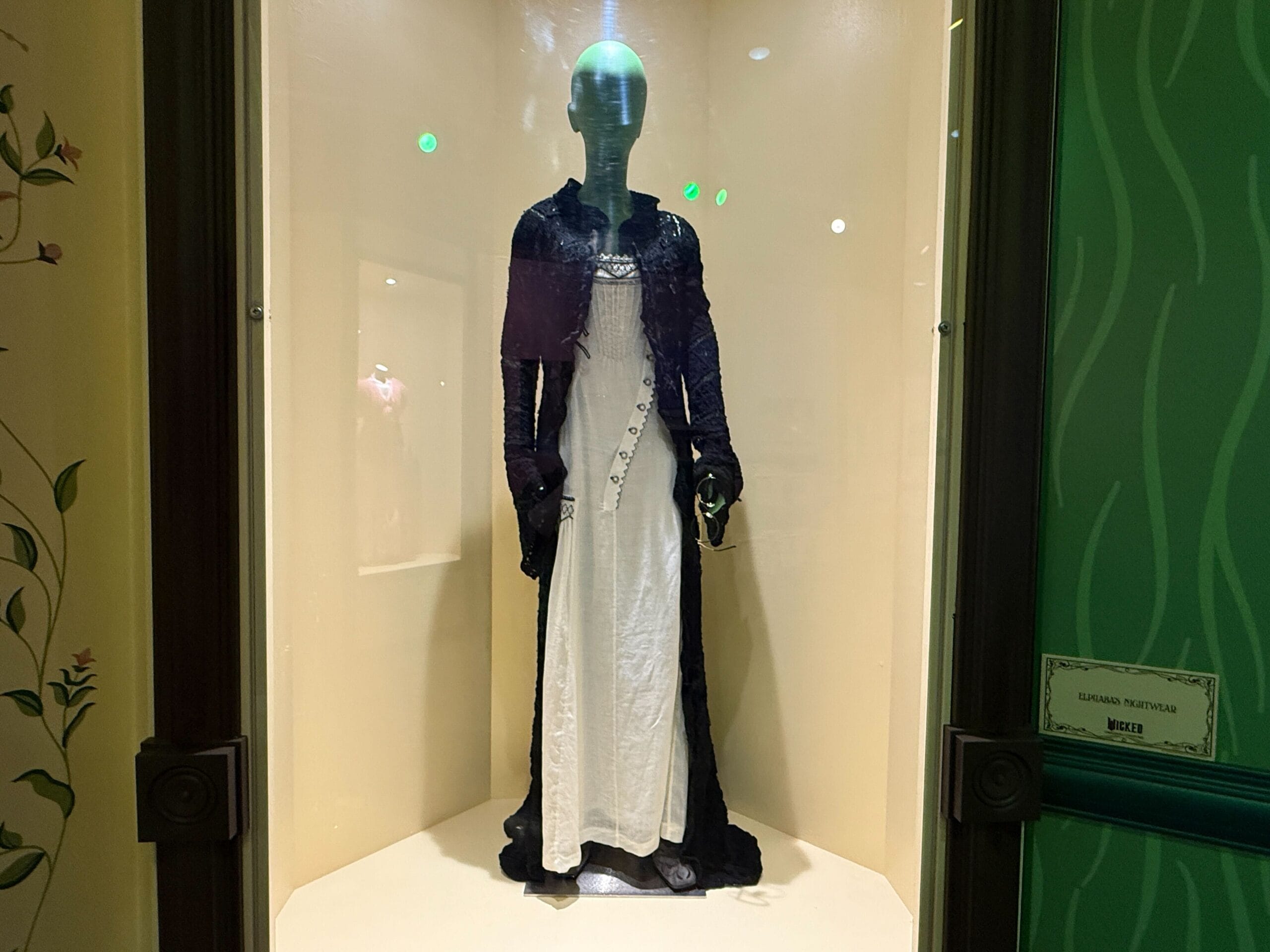 A mannequin exhibits a long white dress with a dark shawl in a glass case against cream walls, similar to Disney parks displays.