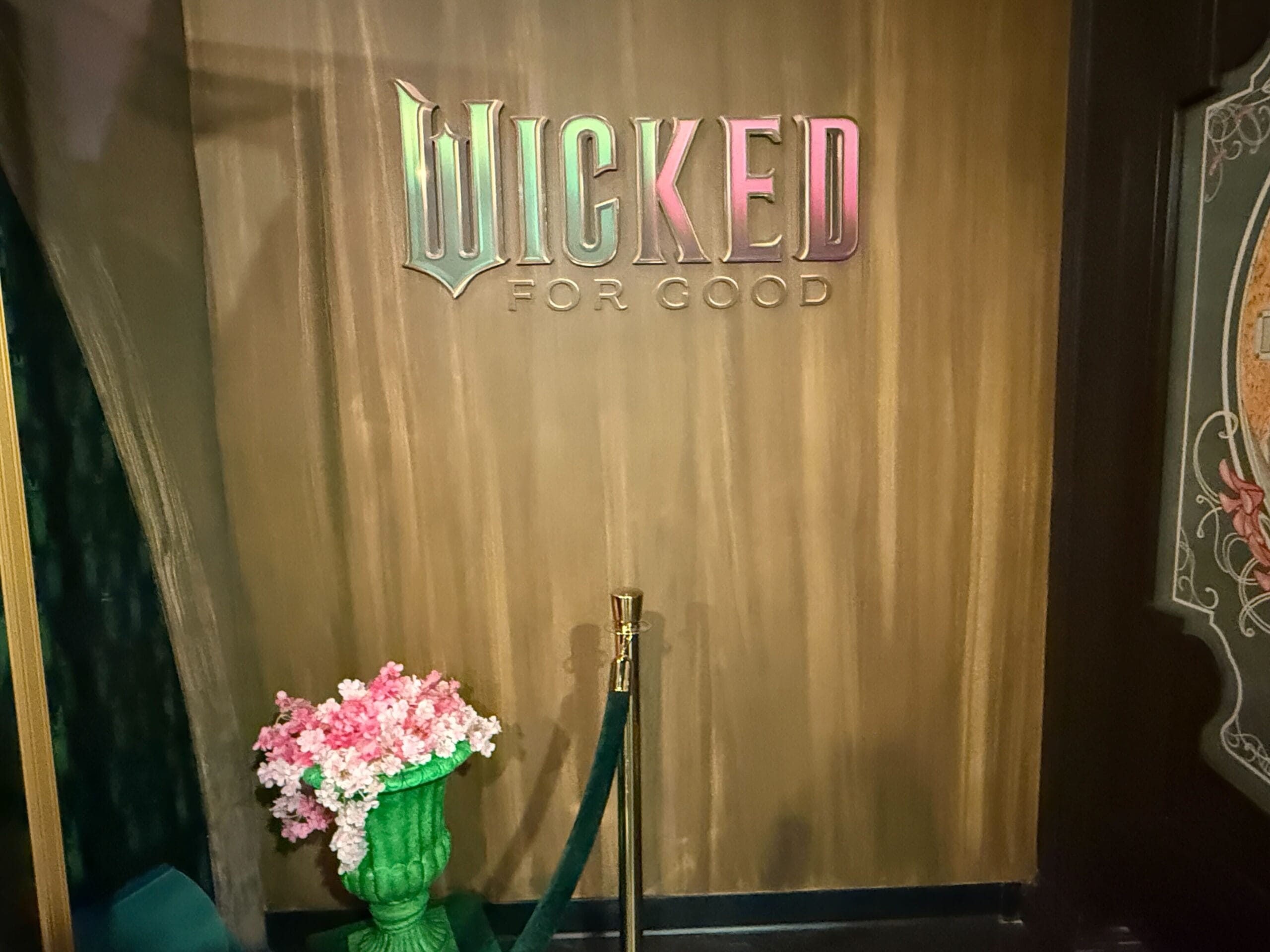 A sign saying "WICKED FOR GOOD" is displayed on a wooden wall above a green vase of pink flowers, reminiscent of Disney decor details.