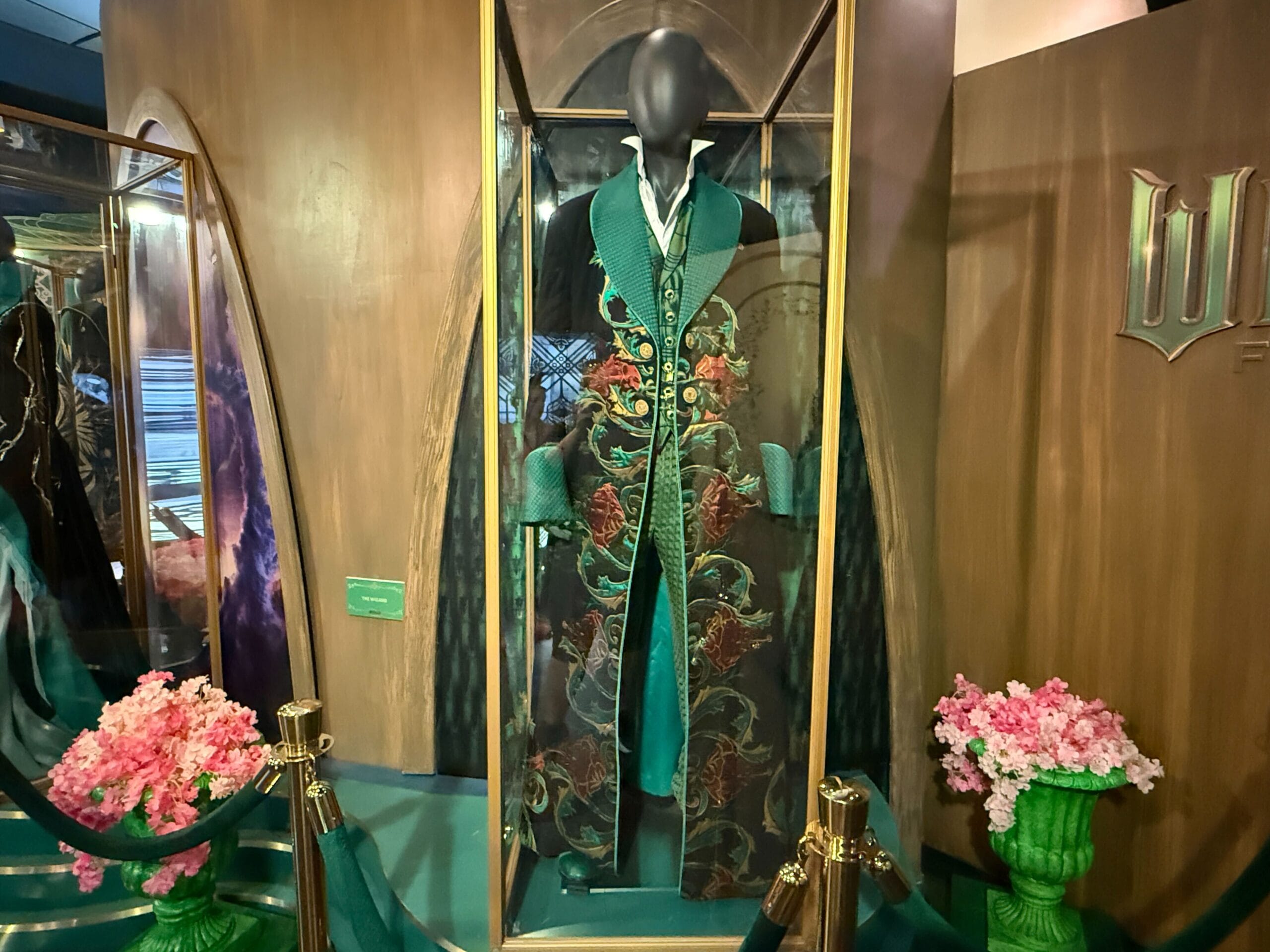 An ornate green and black robe in a glass case, flanked by pink flowers—evoking the detailed displays at Disney Parks.