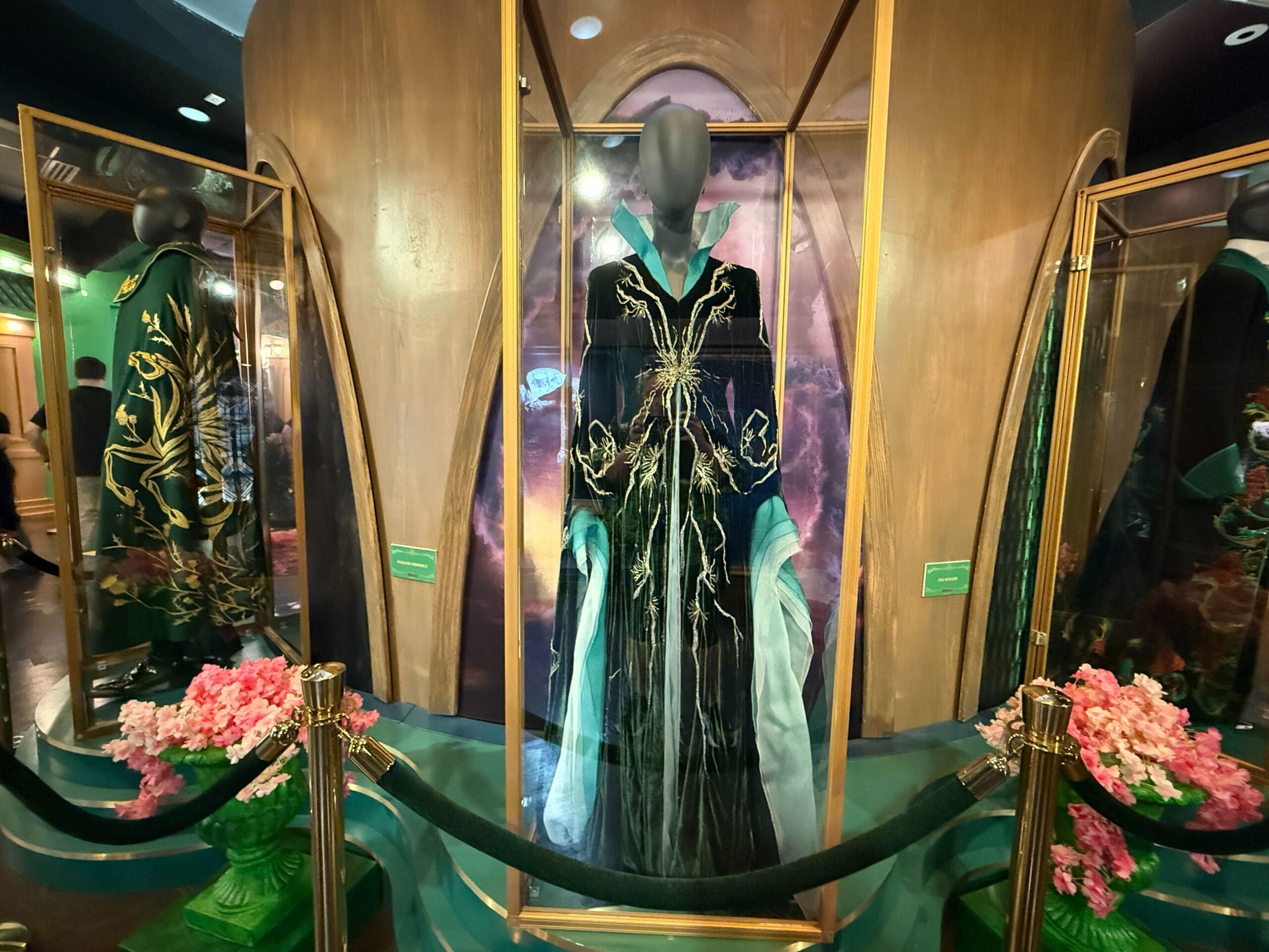 An elegant, gold-embroidered robe is showcased behind glass at Disney, surrounded by floral displays and themed costumes.
