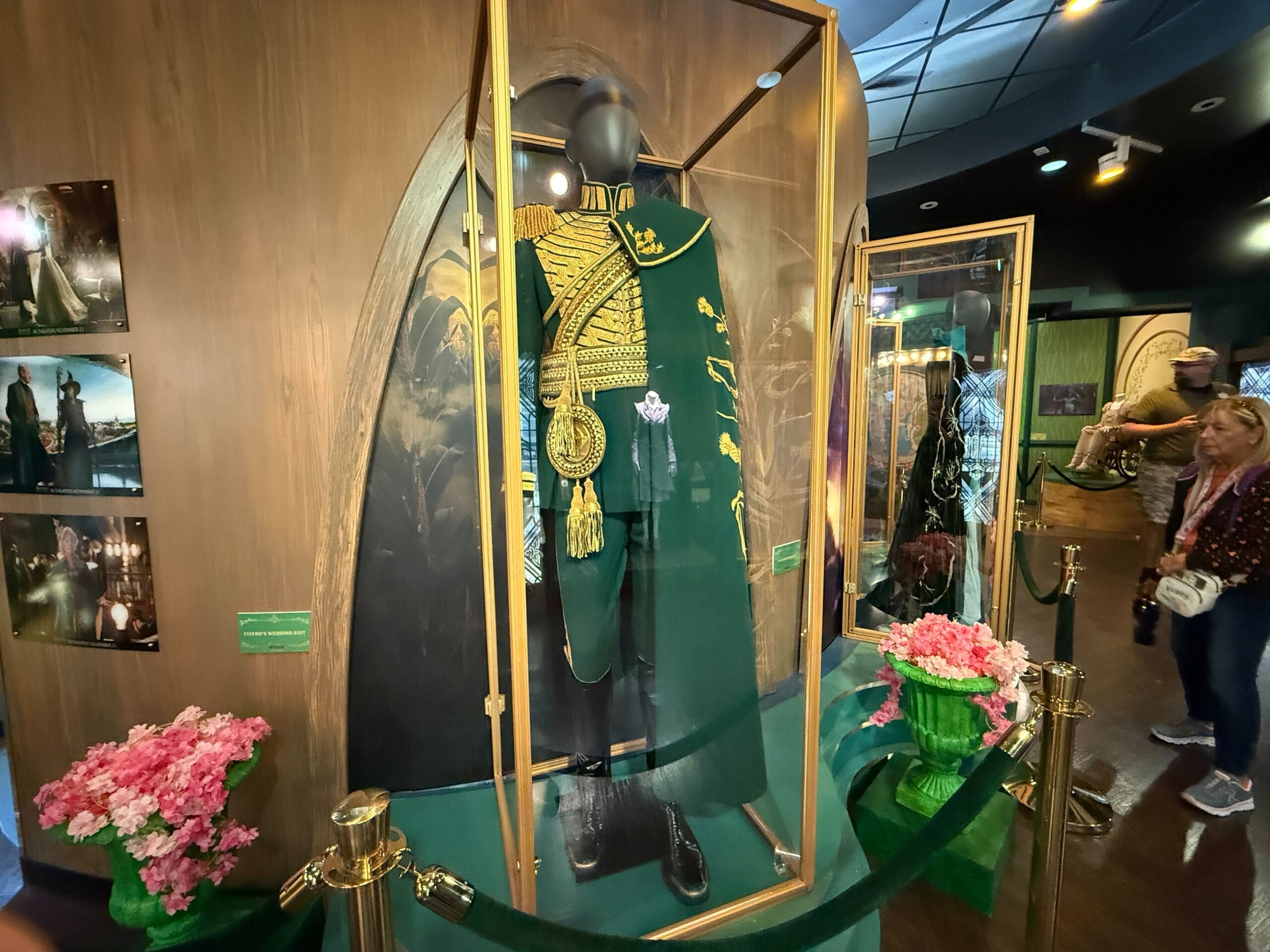 A green, gold-trimmed ceremonial costume is showcased in a glass case with flowers and photos, like an exhibit at a Disney park.