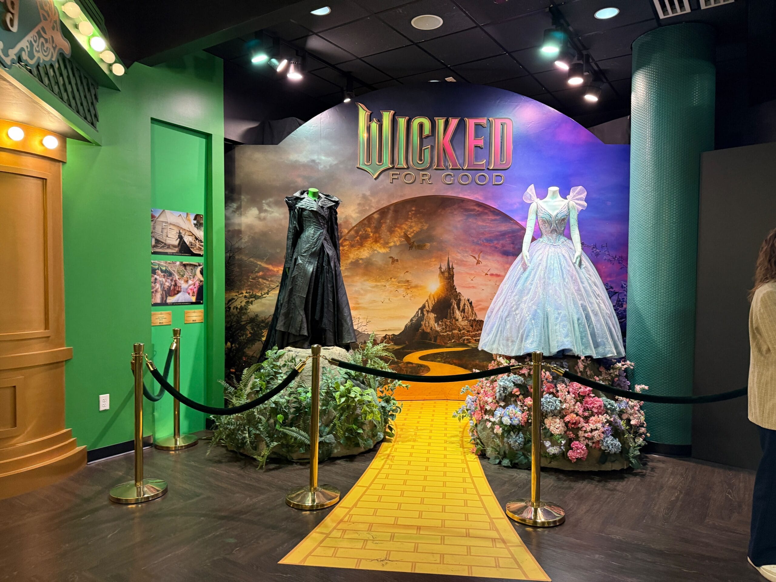Two "Wicked" costumes on mannequins with a yellow brick road, evoking Fantasyland vibes from Disney parks' colorful settings.