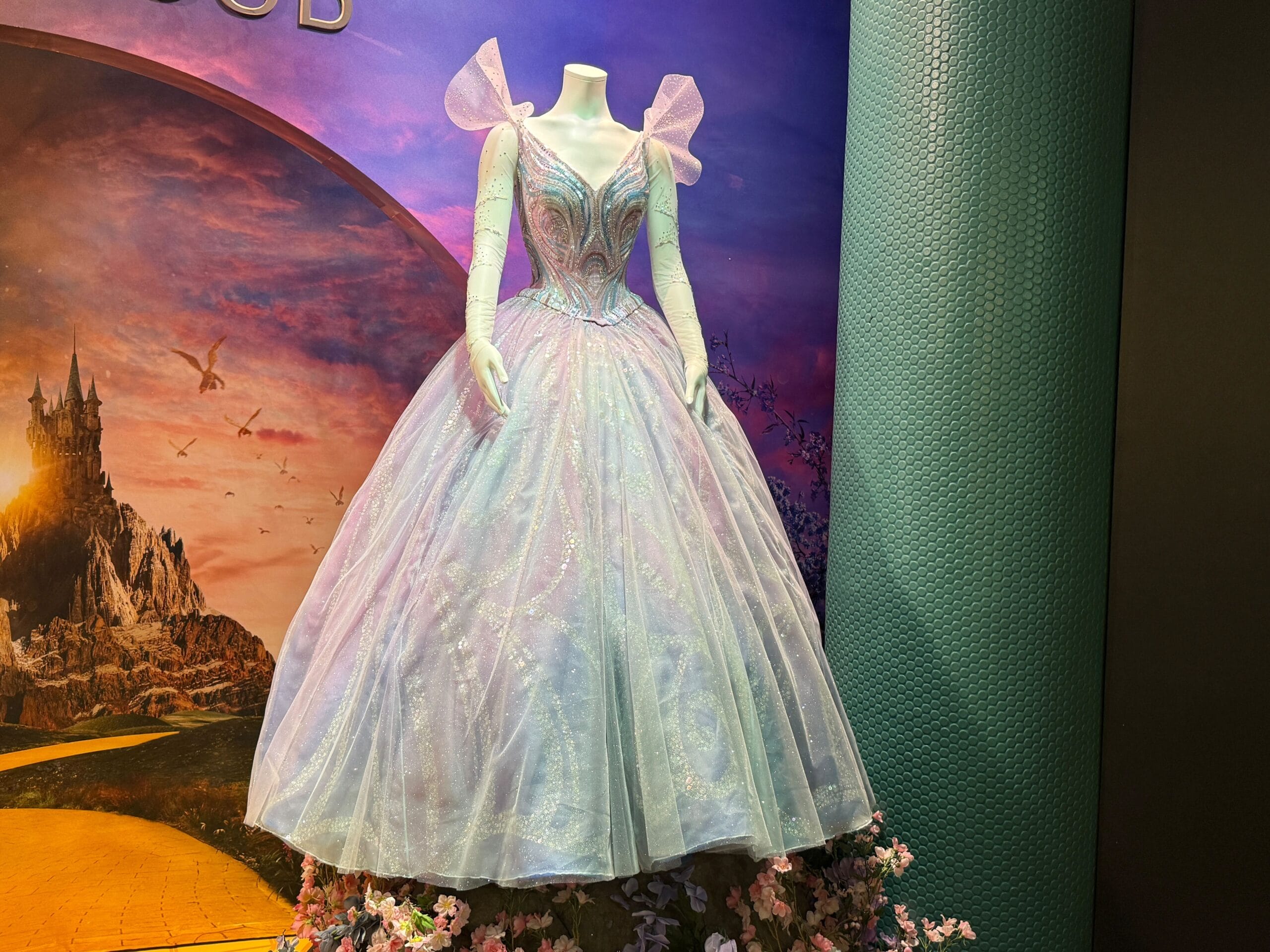 A sparkling, light blue ball gown with sheer sleeves on a mannequin stands before a castle backdrop resembling Disney’s Cinderella Castle.