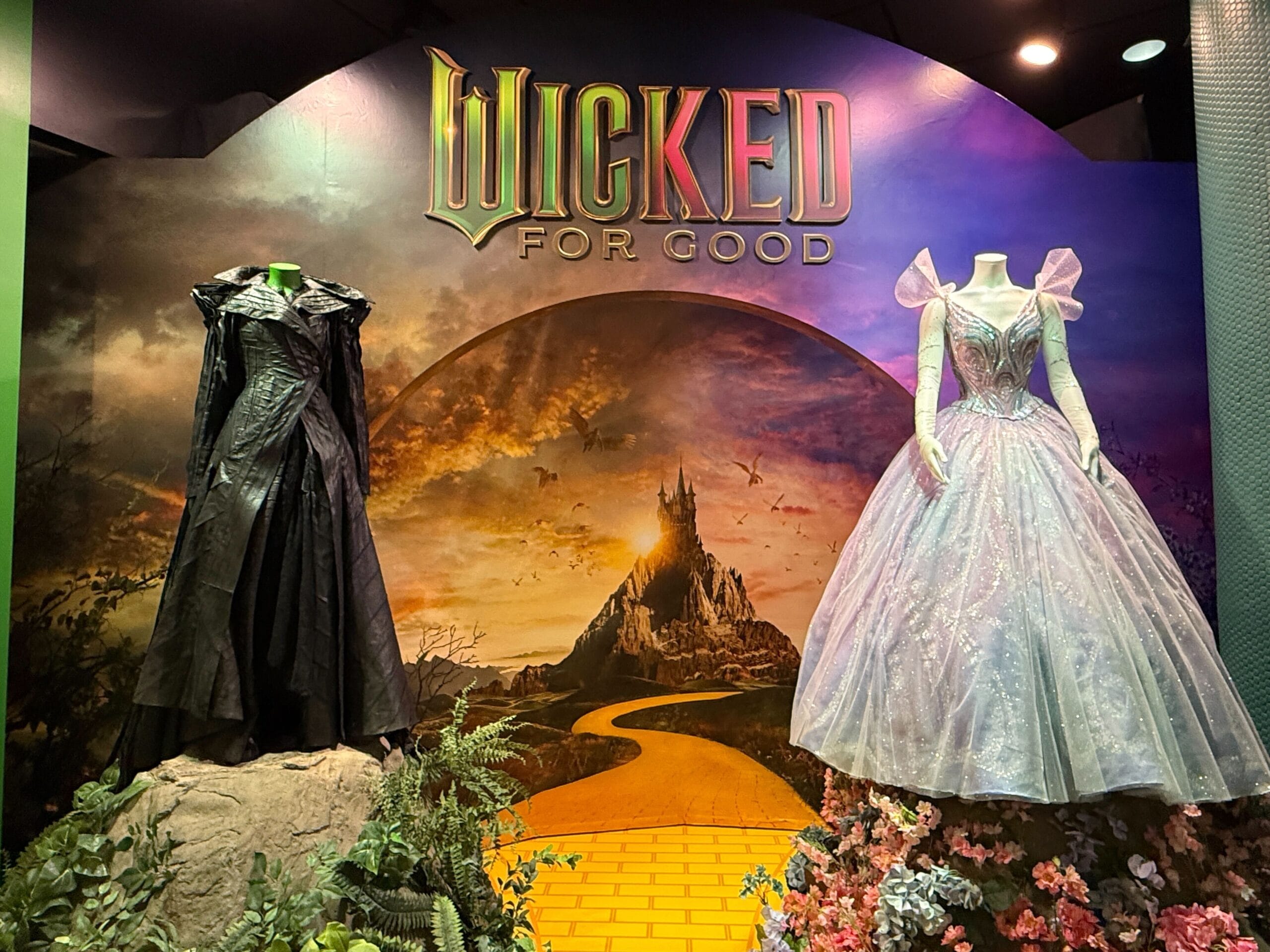 Two mannequins in black and pink gowns stand before a "Wicked For Good" sign, yellow brick road, and a castle not like Disney’s castles.