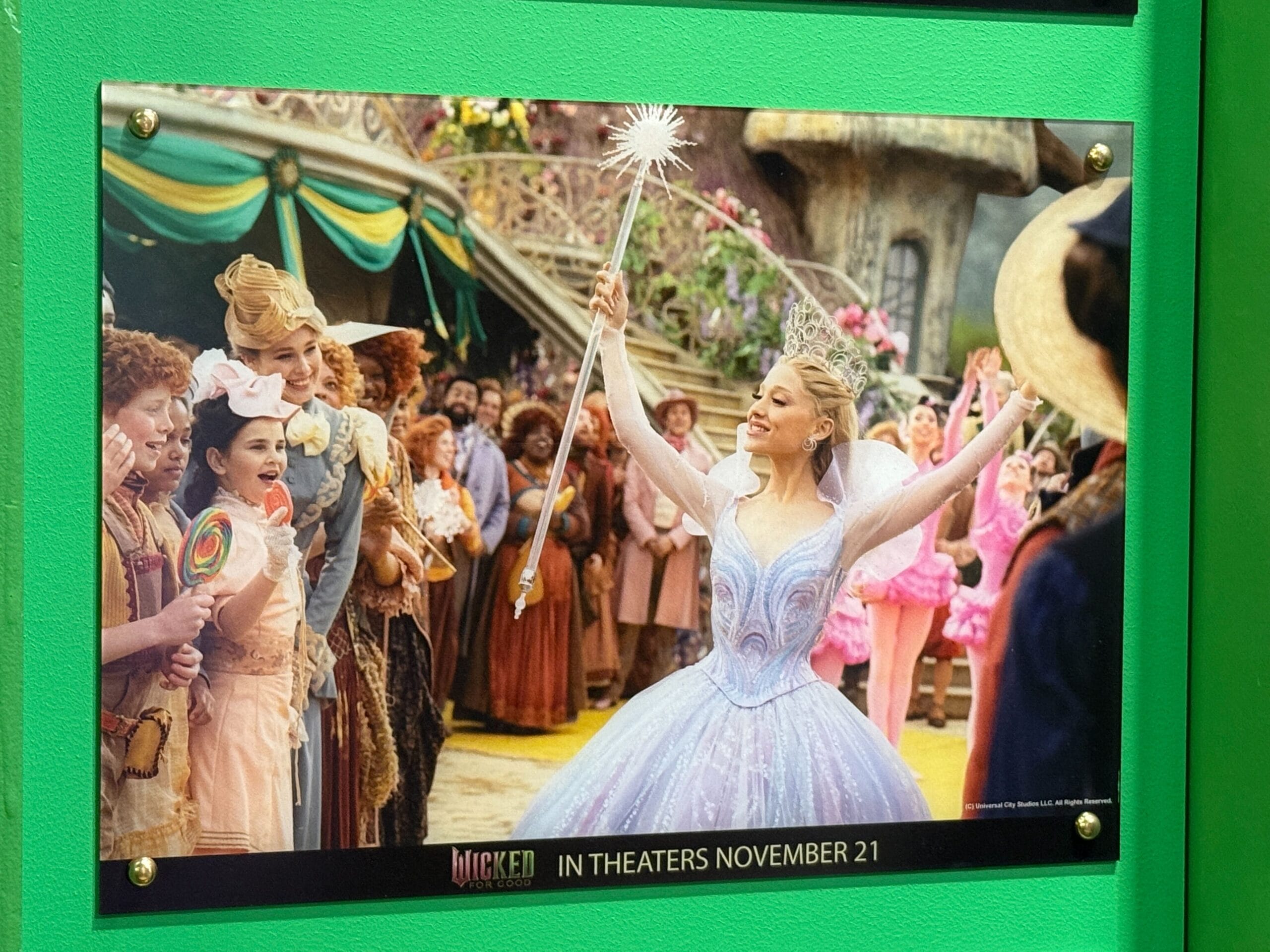 A woman in a sparkling gown holds a wand, surrounded by people in colorful costumes at a Disney park’s outdoor celebration.