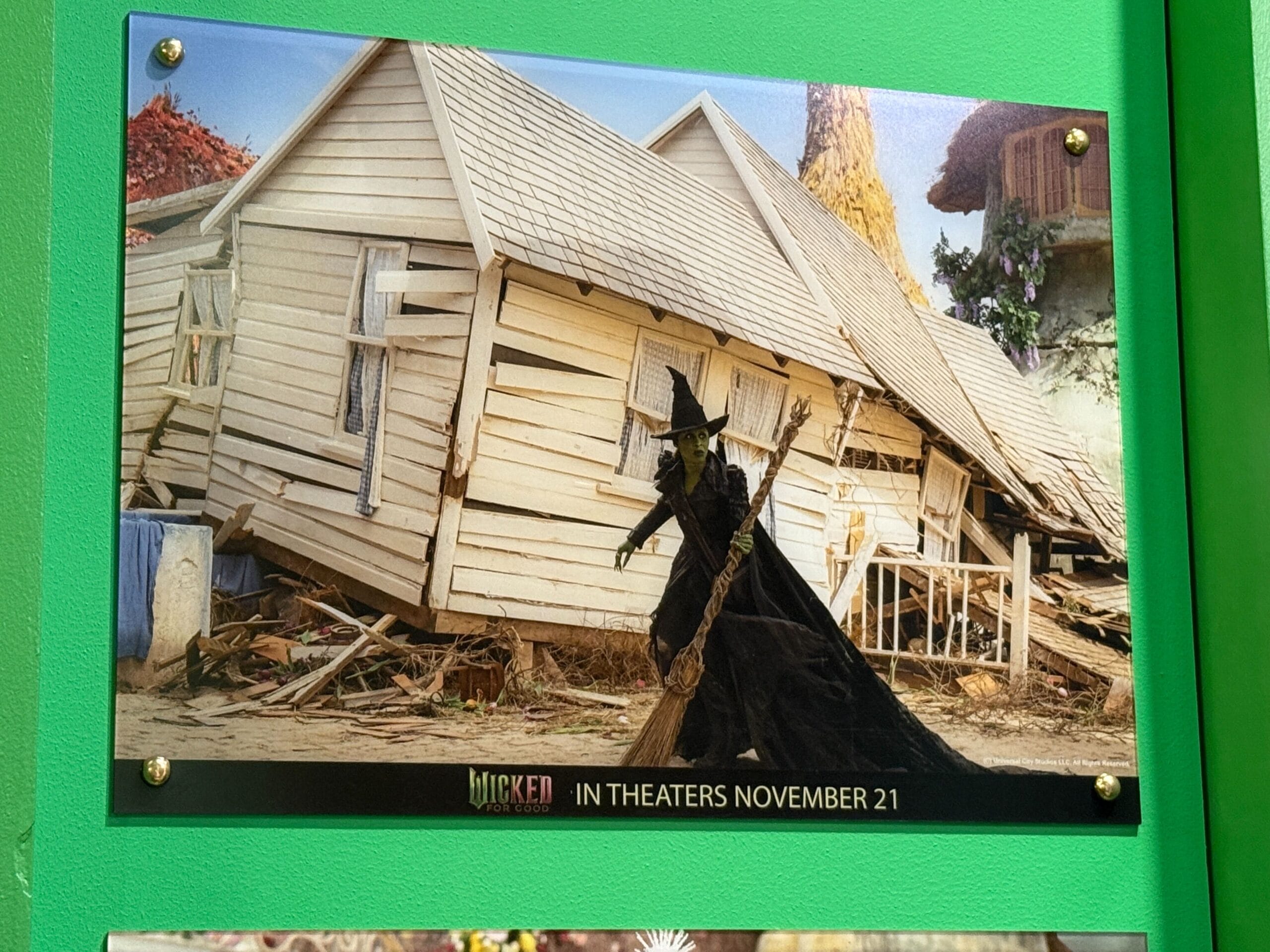 A green-skinned witch with a broom stands before a collapsed white house, not resembling any Disney castle. Text: “Wicked, In Theaters Nov 21.”