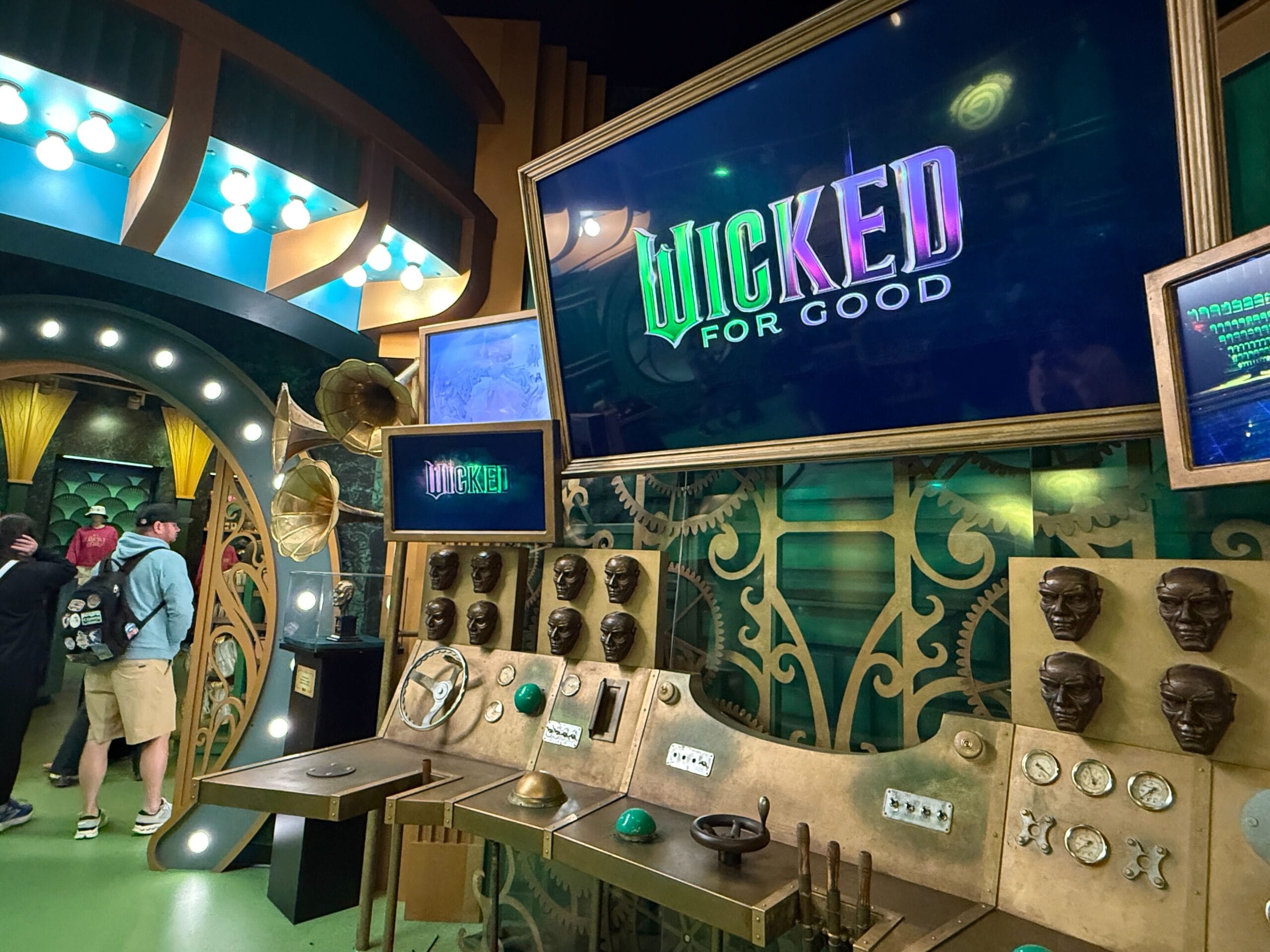 An emerald-hued room with "Wicked for Good" on a screen, vintage controls, and guests exploring like an immersive Disney exhibit.