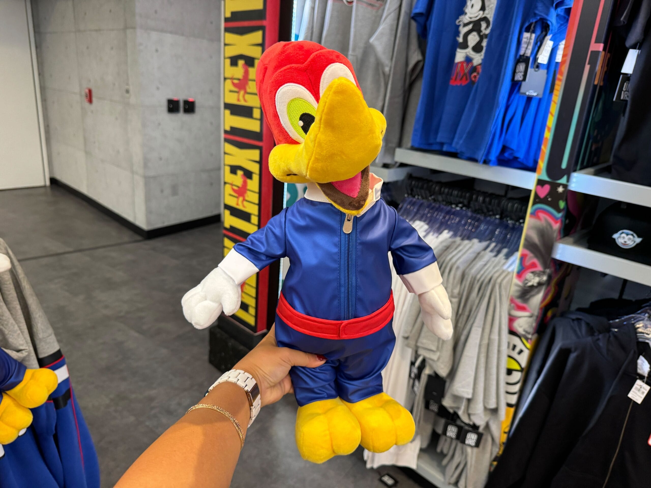 A hand holds a Woody Woodpecker plush toy wearing a blue jumpsuit in a clothing store.