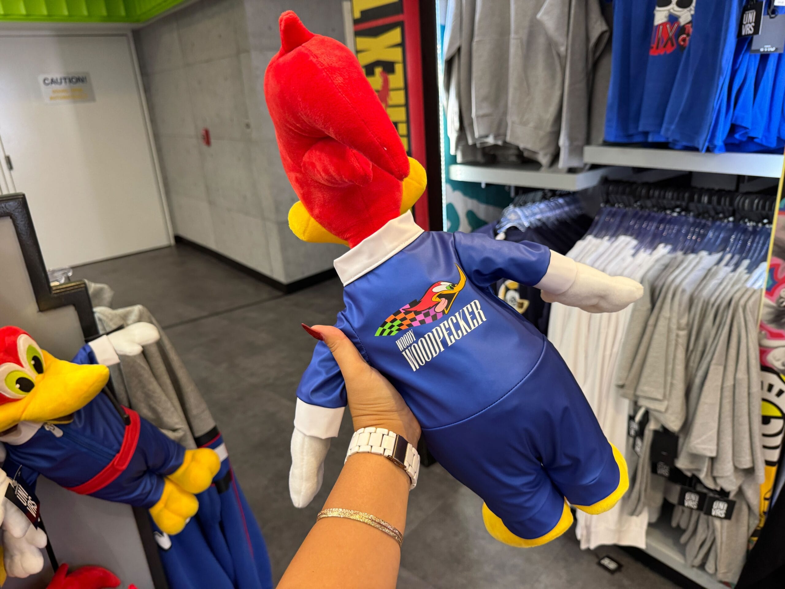 A hand holds a Woody Woodpecker plush in a blue racing suit inside a clothing store.