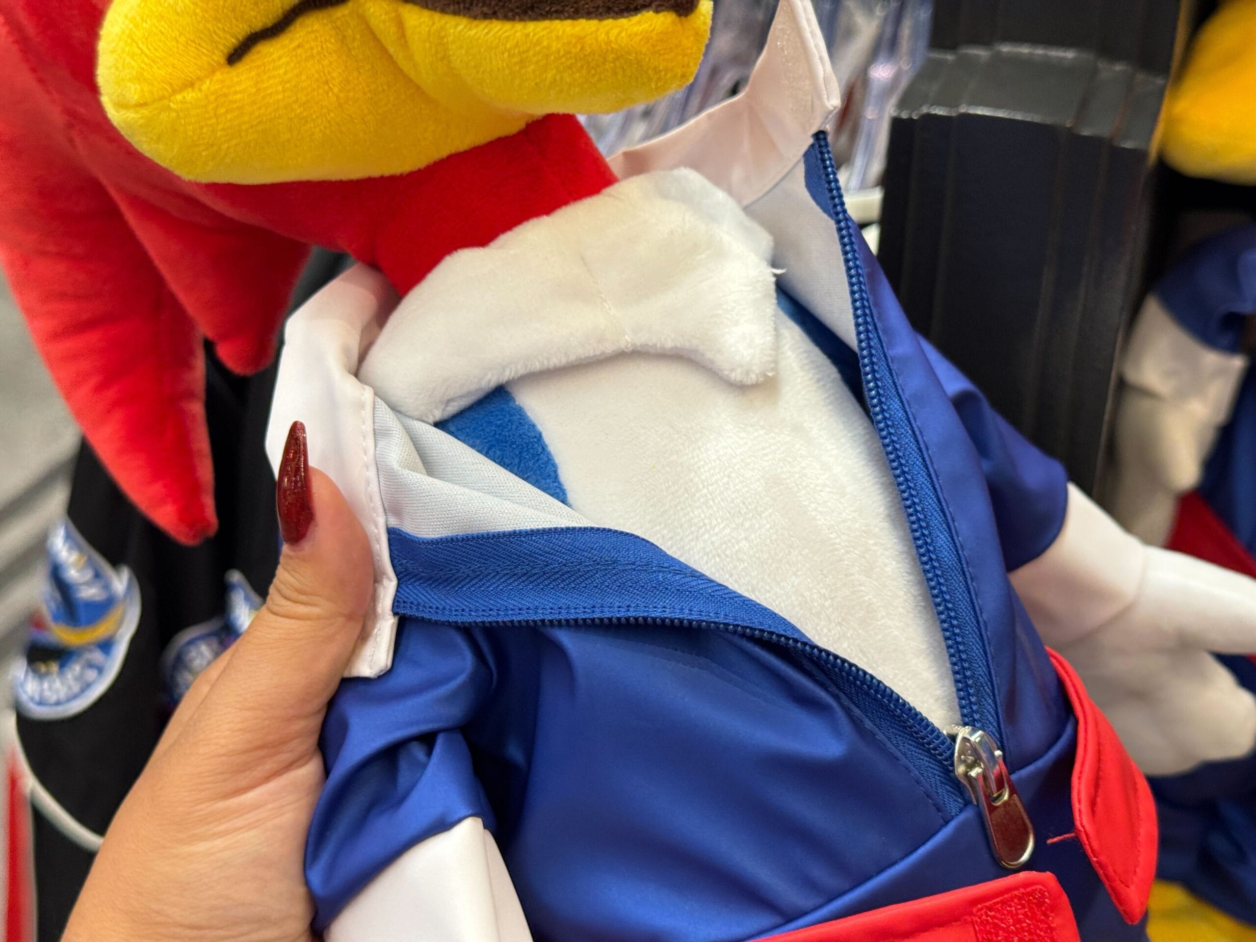 A hand unzips a blue and white jacket on a red plush toy, revealing a hidden blue triangle on its chest.