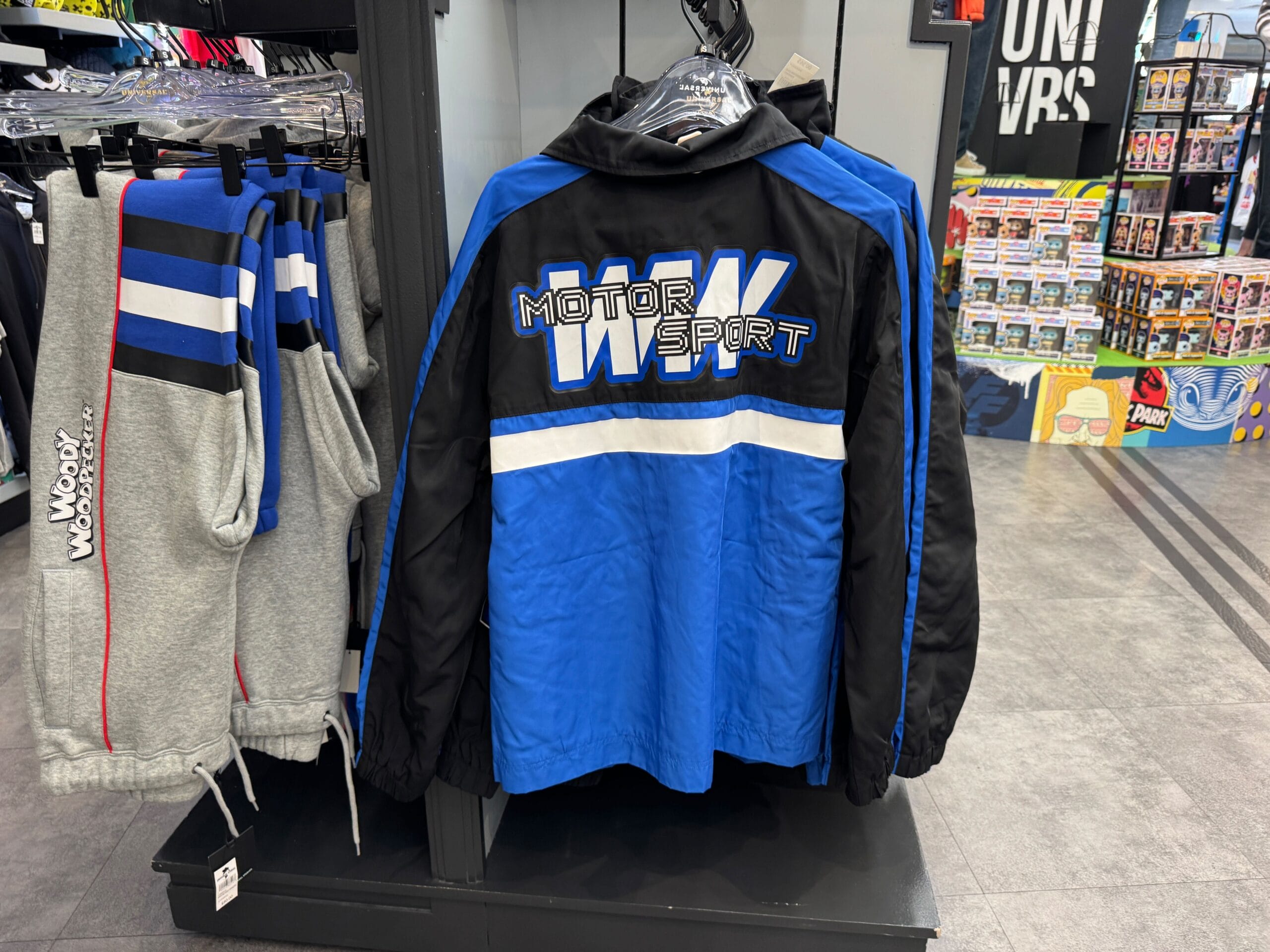 A blue and black "MOTOR SPORT" jacket hangs on a rack next to gray sweatpants in a retail store.