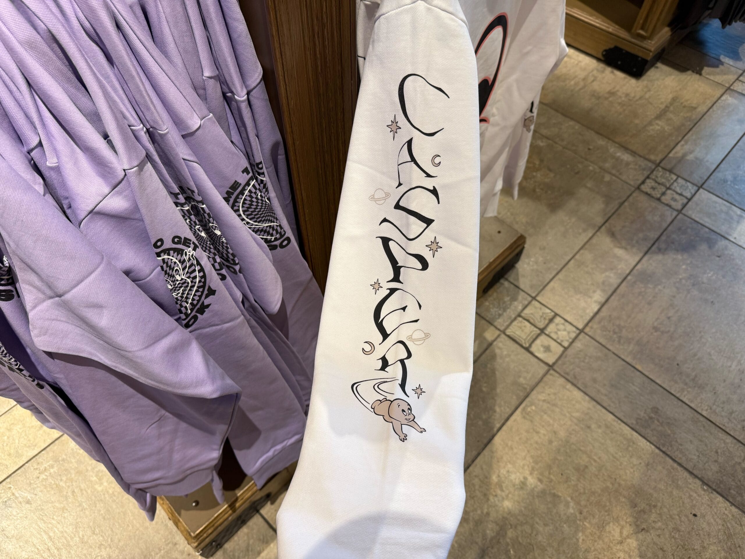 White sleeve reading “Canada” with a cartoon moose, similar to Epcot Canada pavilion items; lavender shirts like Disney merch nearby.