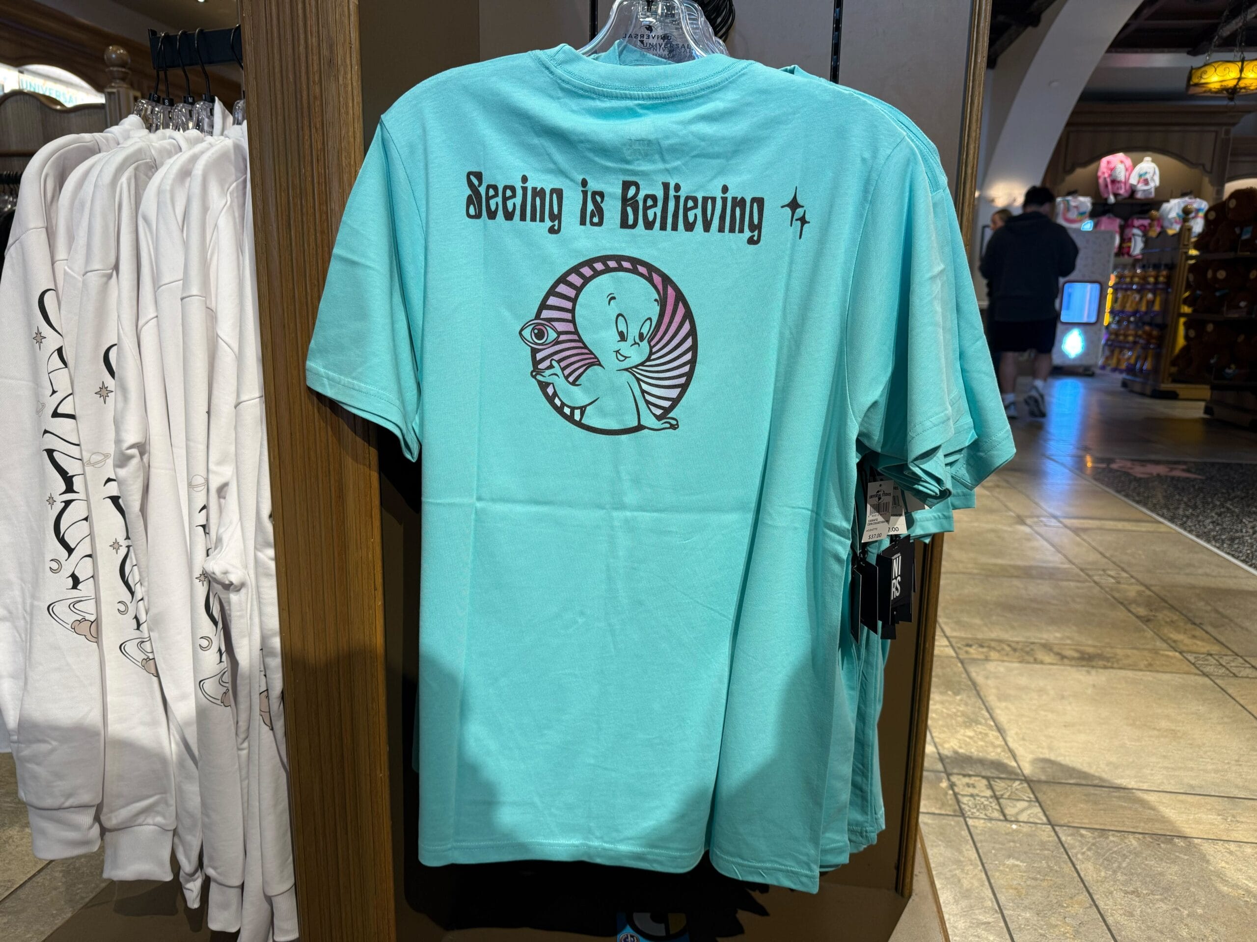 A turquoise t-shirt featuring a cartoon ghost and “Seeing is Believing,” similar to playful Disney park merch but no castle shown.