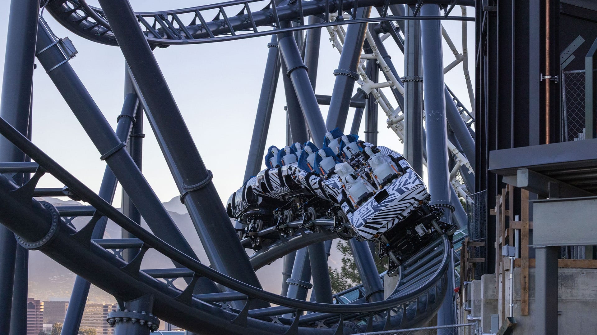 A zebra-striped roller coaster train races around a sharp turn on steel tracks, with scenic mountains like those in Disney parks behind.