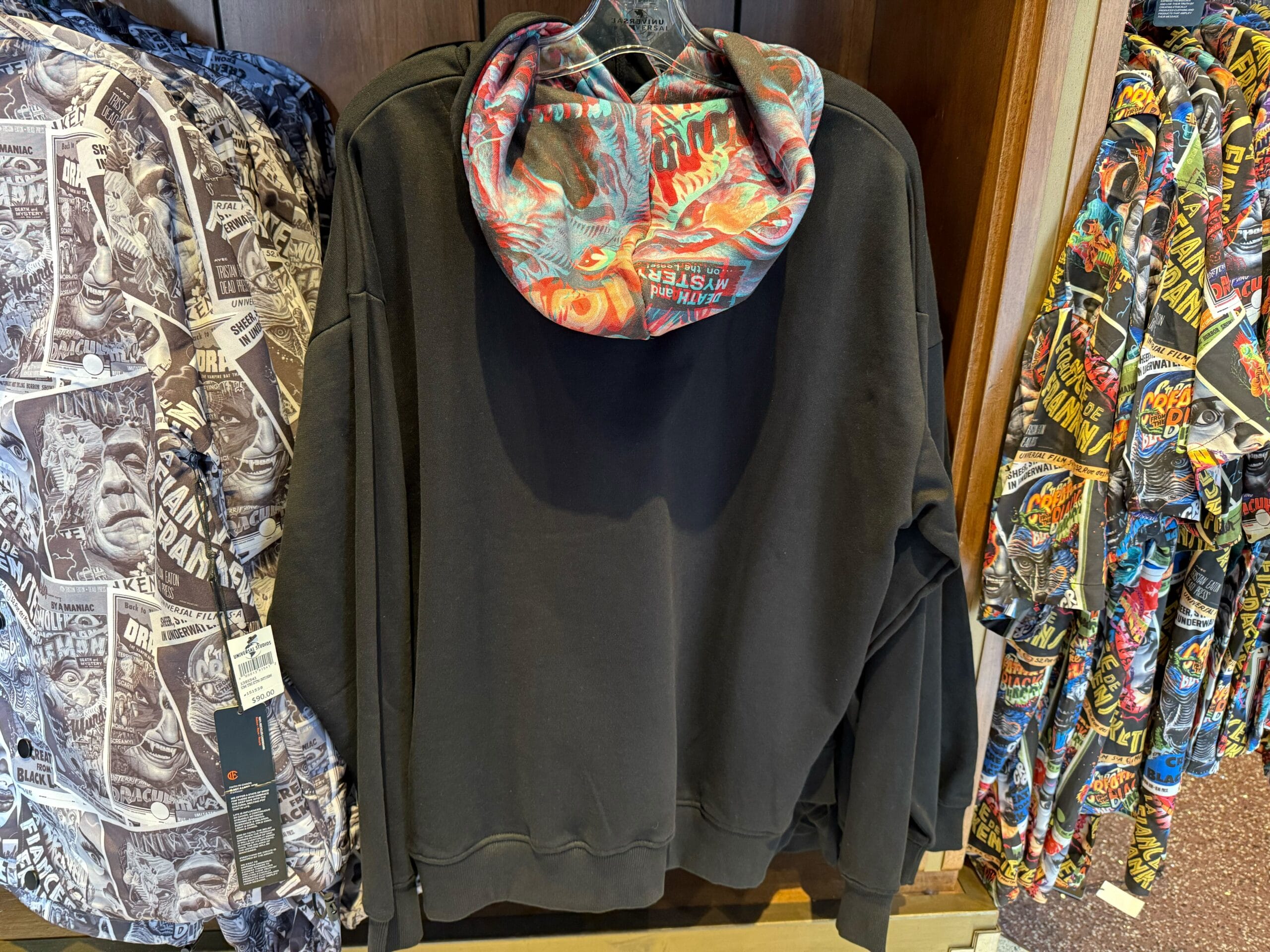 Black hoodie featuring a colorful comic-style hood from the Universal Monsters Collection, displayed on a rack—not Disney Parks merch.