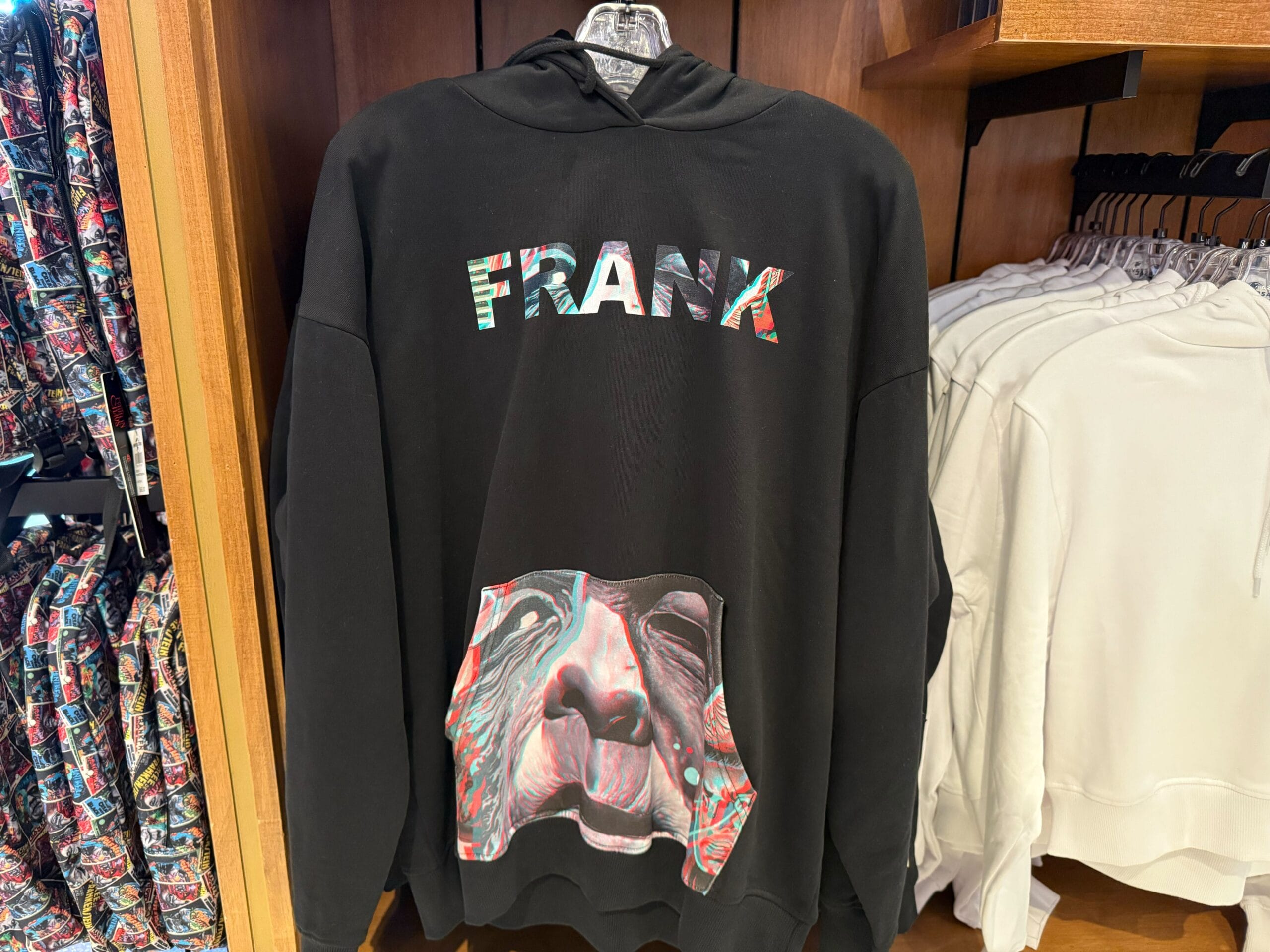 A black hoodie with "FRANK" in bold, a colorful sloth face, and retro monster style, seen hanging on a store rack—not Disney merch.
