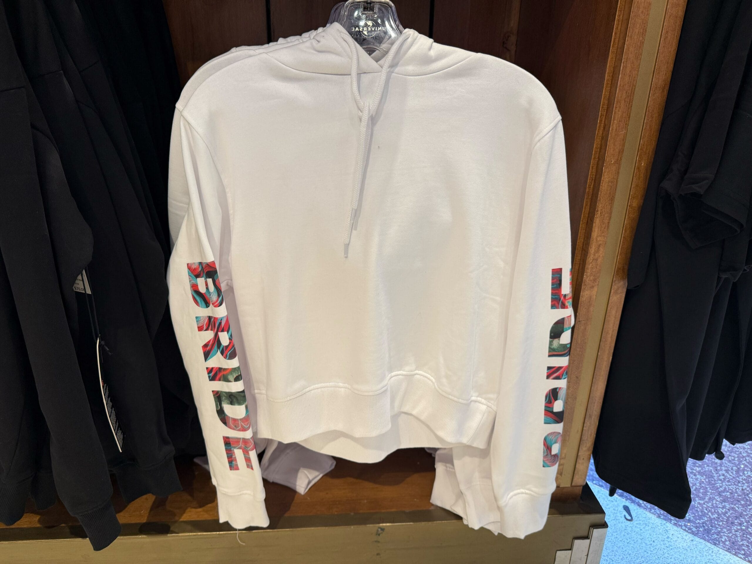 White hoodie from the Universal Monsters Collection with "PRIDE" in vibrant letters, set on wood—no castle like at Disney Parks.