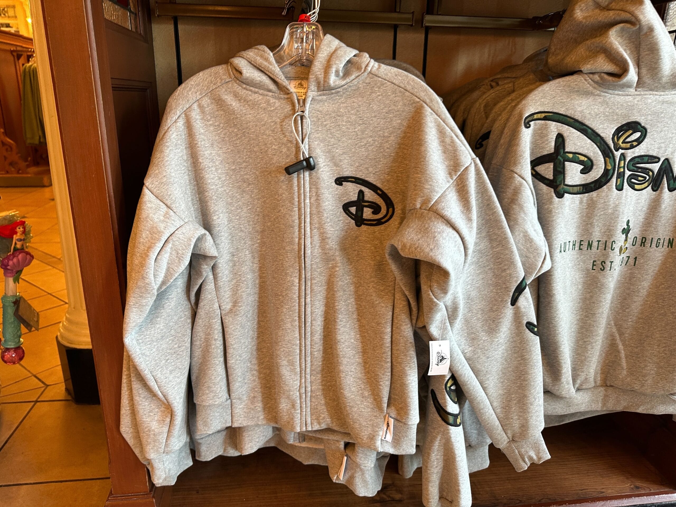 Gray zip-up Disney hoodie with a small chest logo, hanging in a park shop—just steps from one of the famed Disney castles worldwide.