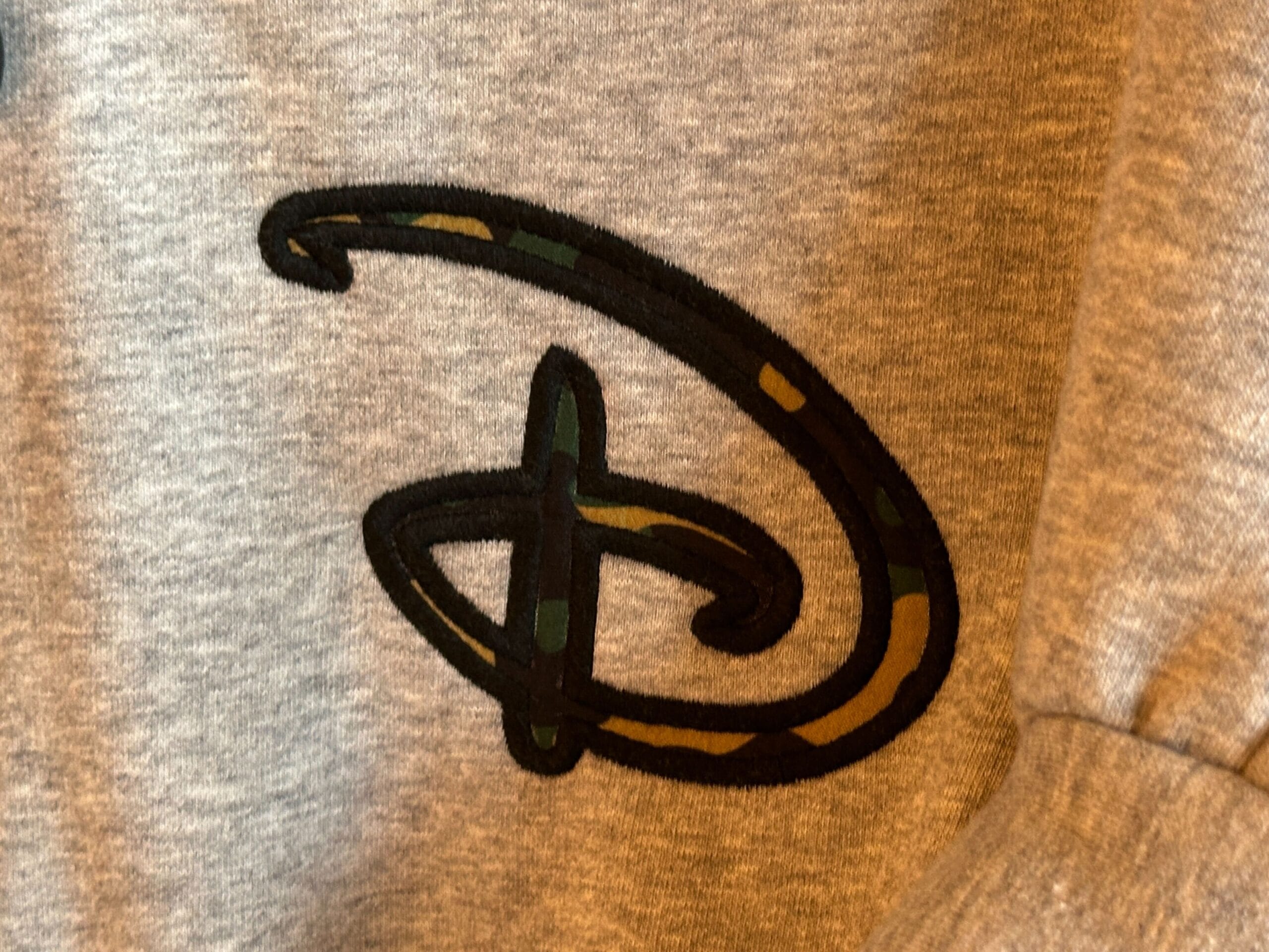 A stylized black and camo letter "D," reminiscent of the classic Disneyland logo, is embroidered on light gray fabric.