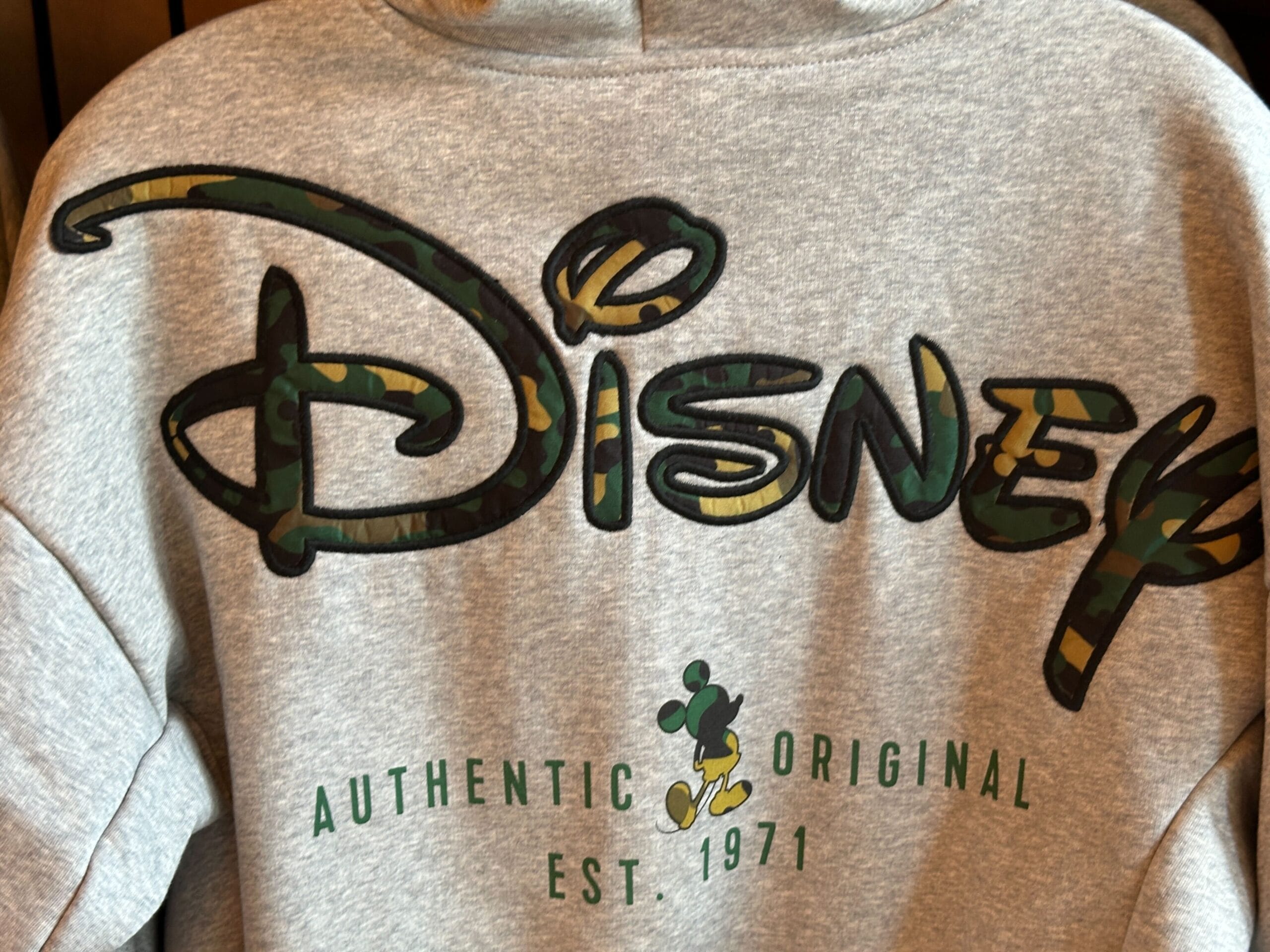 Gray sweatshirt featuring camo "Disney" logo and small Mickey Mouse, with "Authentic Original Est. 1971" in green, evoking park style.