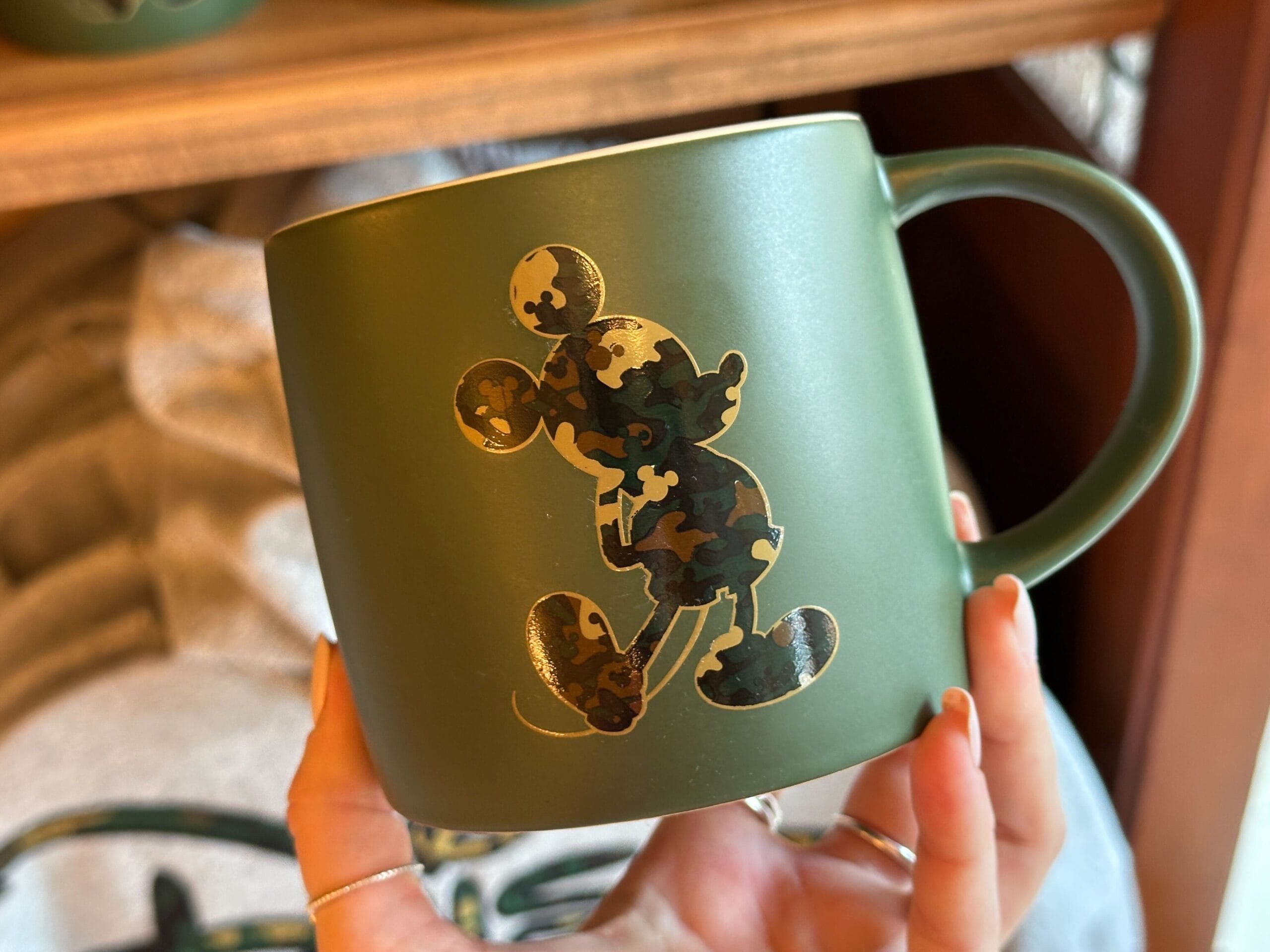 A green mug featuring a camouflage-style silhouette of a mouse character, reminiscent of classic Disney icons from the theme parks.