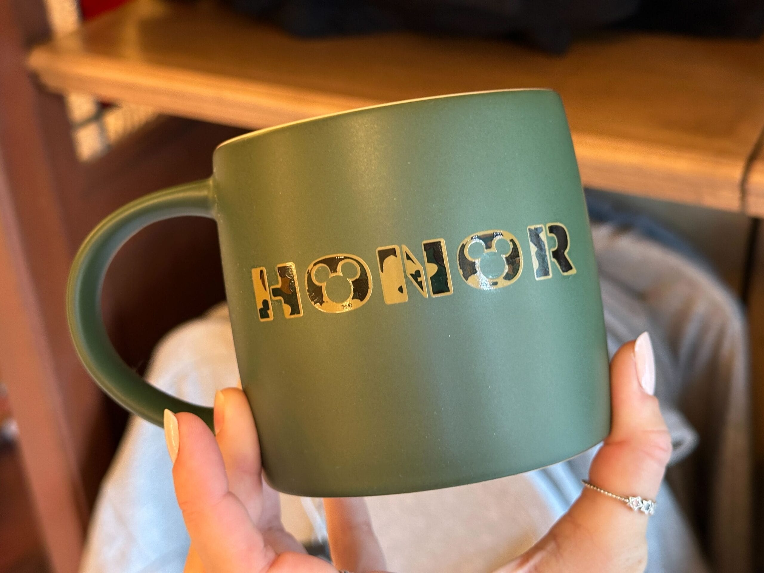 A green mug reading "HONOR" in stencil letters, held by someone with pale nails, perfect for sipping at a Disney park café.