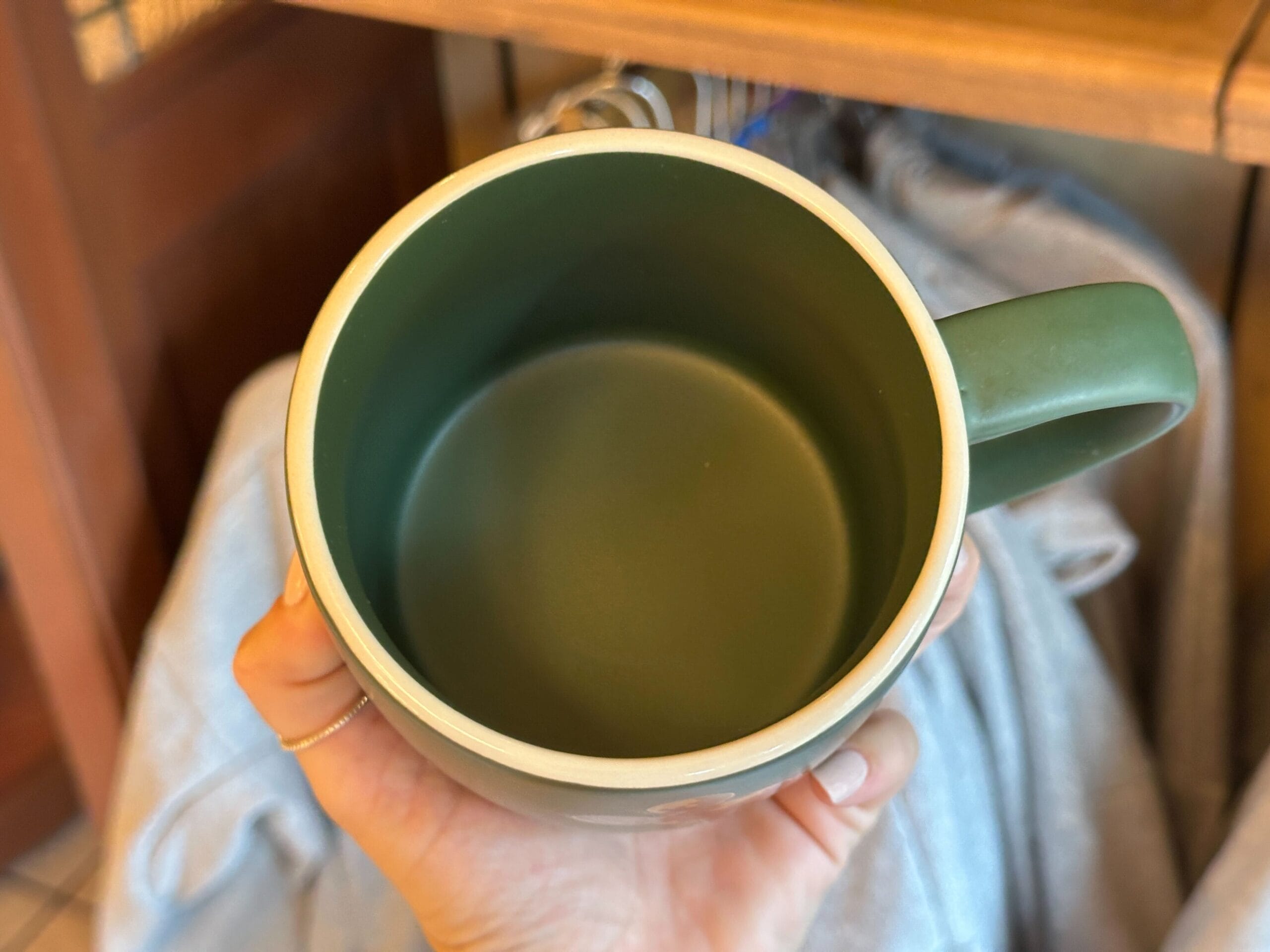 A hand holding an empty green mug above a soft gray fabric and wooden shelves, reminiscent of cozy Disney resort decor.