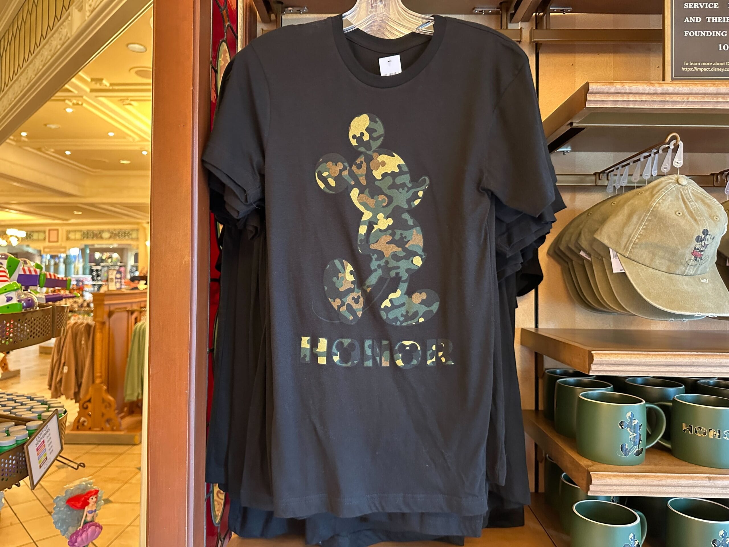 Black t-shirt featuring a camo Mickey Mouse silhouette and "HONOR" text, sold in a Disney park shop, no castle in this image.