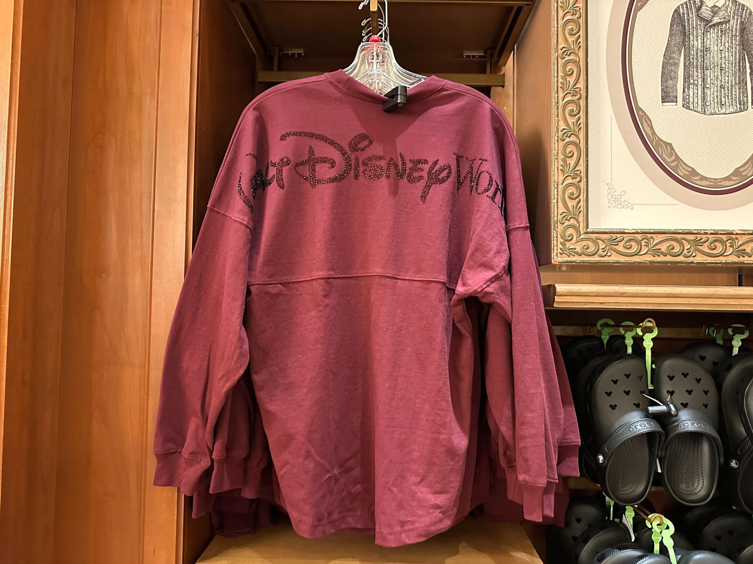 A maroon long-sleeve shirt with "Walt Disney World" in black text, hanging on a wooden rack, referencing the Florida park.