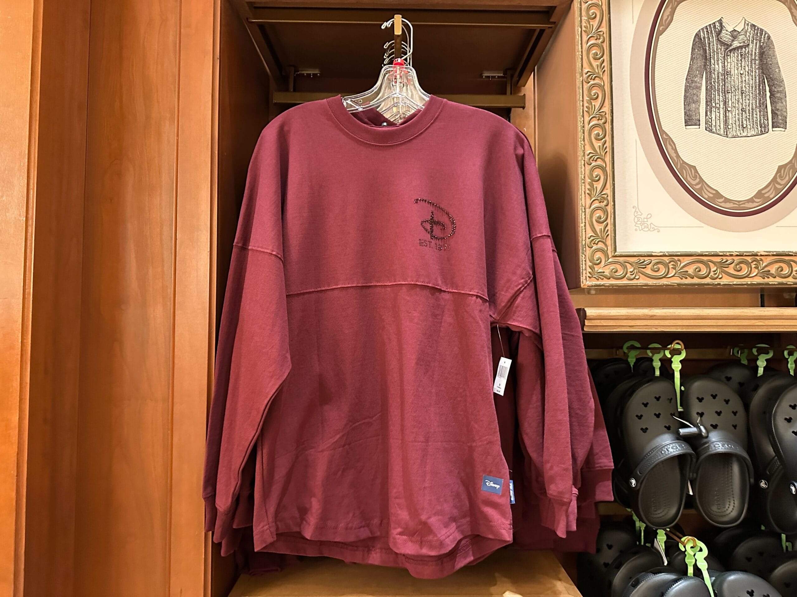 Maroon Disney long-sleeve shirt on rack beside black Crocs in a wooden display, typical of Walt Disney Parks merchandise shops.