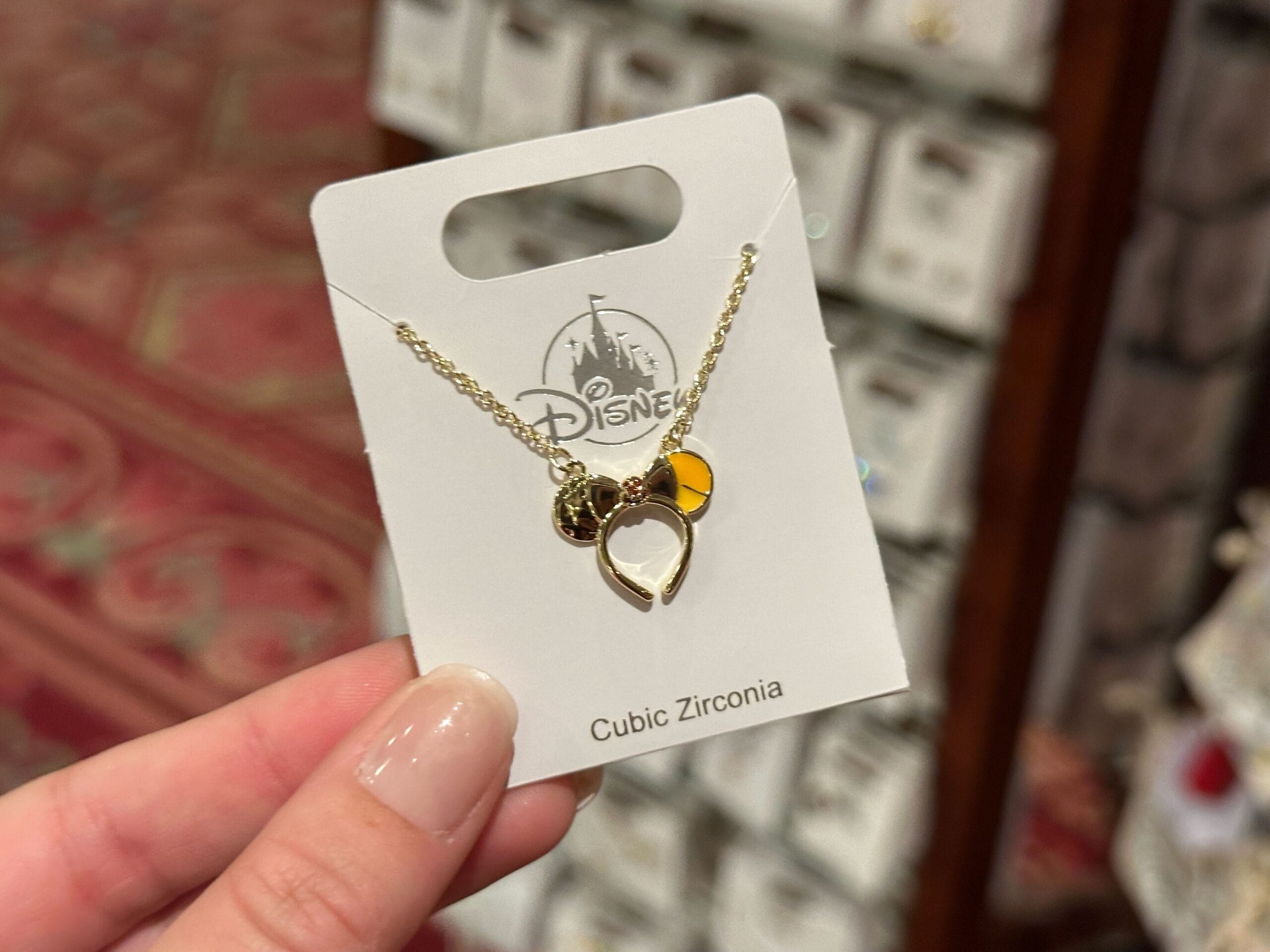 A hand holds a Disney Princess necklace with a Mickey Mouse pendant on a "Cubic Zirconia" card, likely sold at a Disney park.