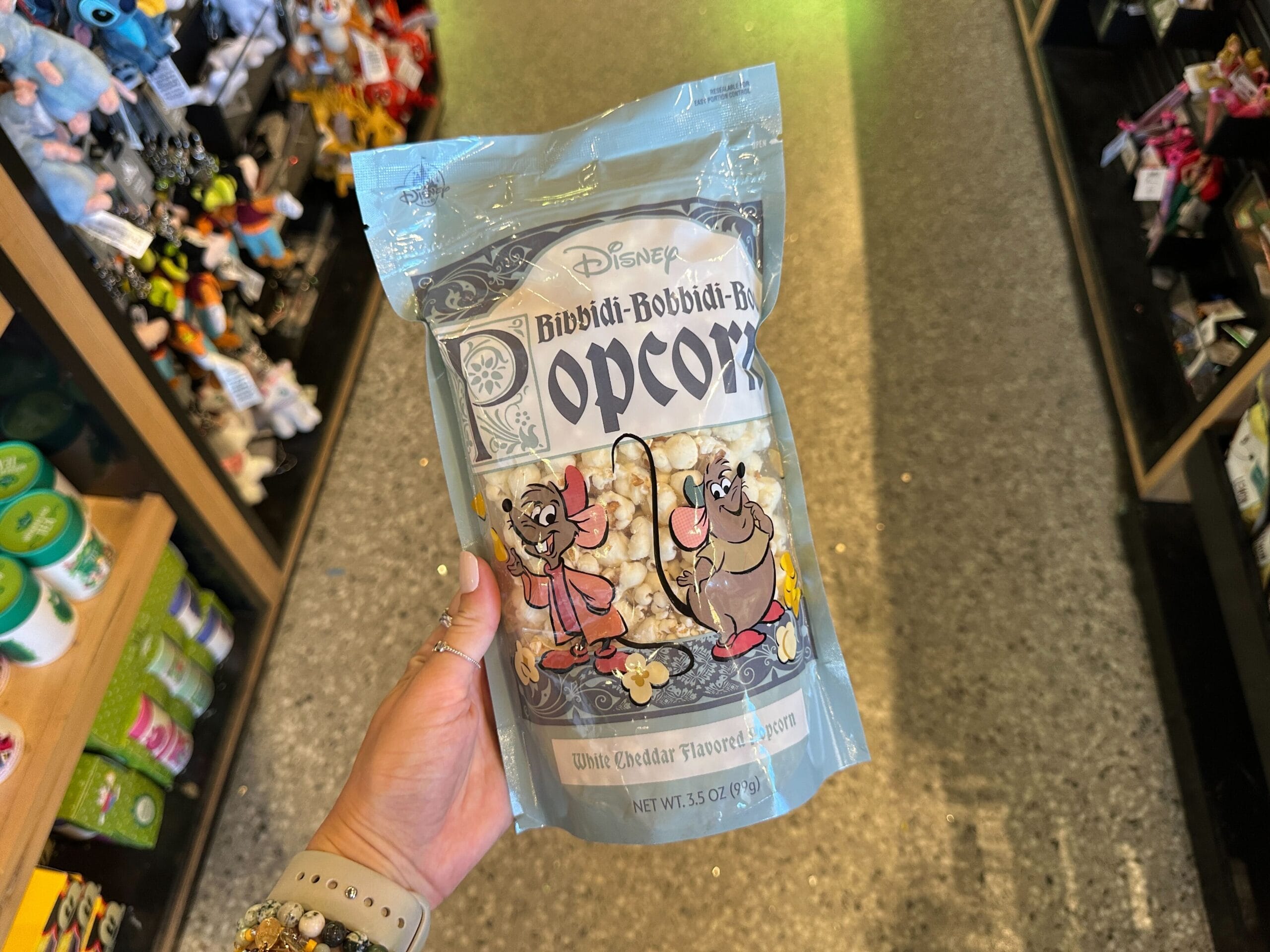 A hand holds Disney Bibbidi-Bobbidi-Boo White Cheddar Popcorn, spotted in a store that could be within a Disney park merchandise shop.
