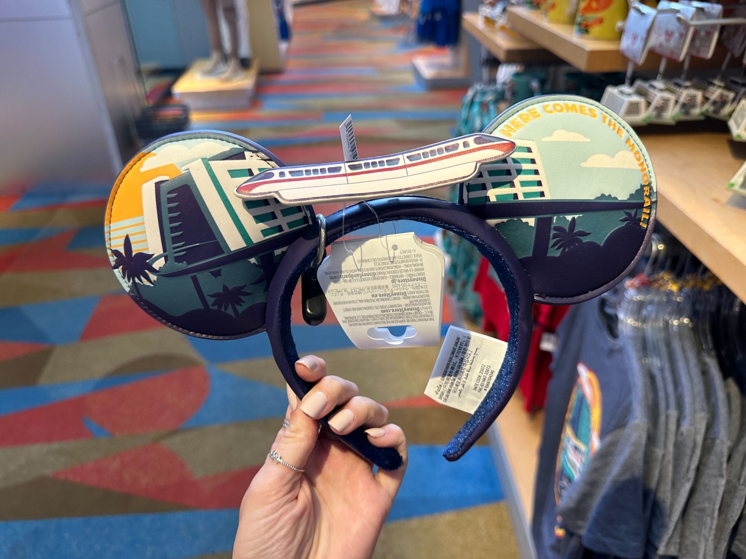 Hand holding Disney ear headband with monorail and sunset motif, displayed in a shop, reminiscent of Walt Disney World themes.