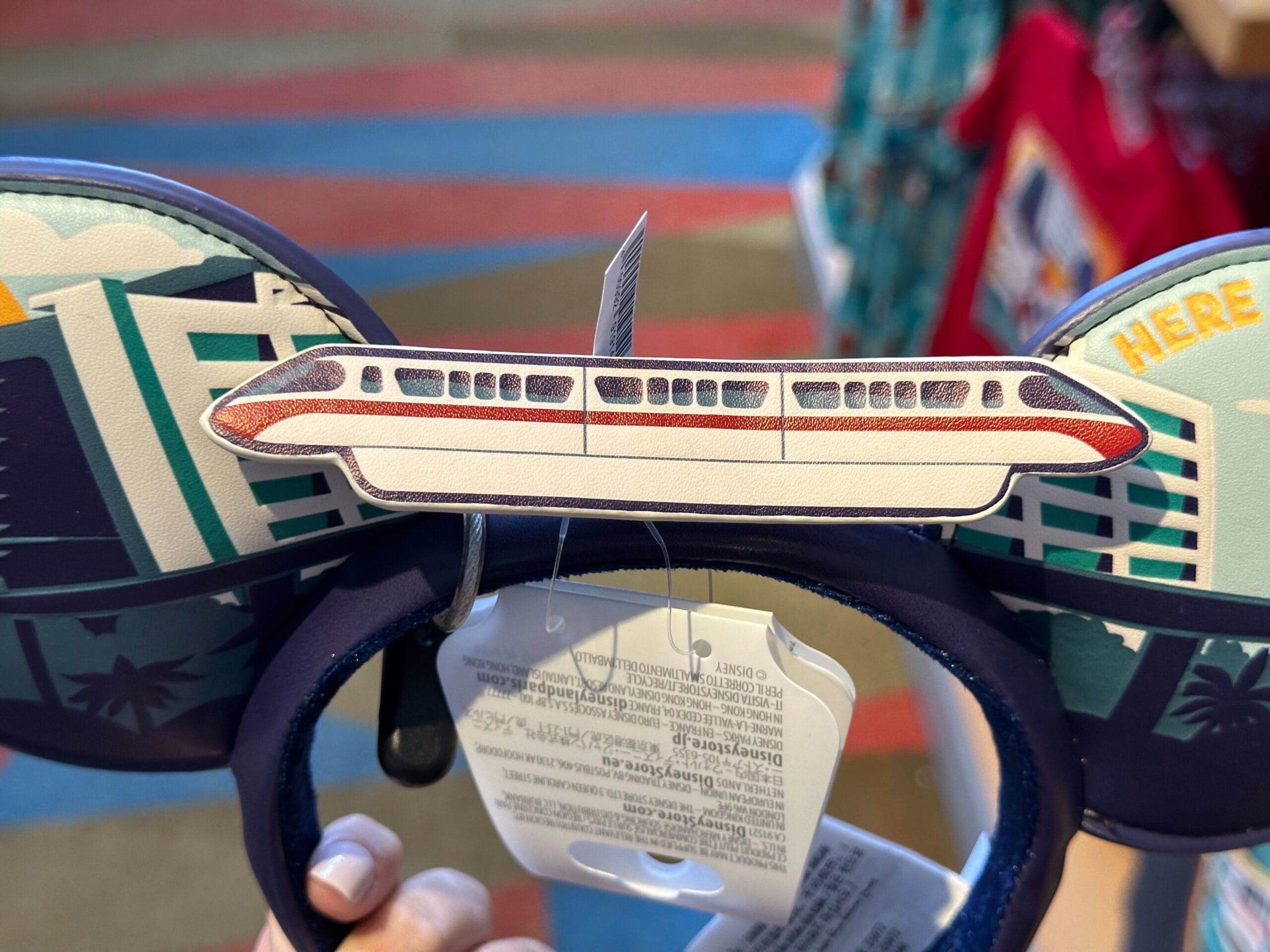 Mickey Mouse ears headband featuring a monorail and cityscape, reminiscent of Disney parks, held by a hand in a shop.