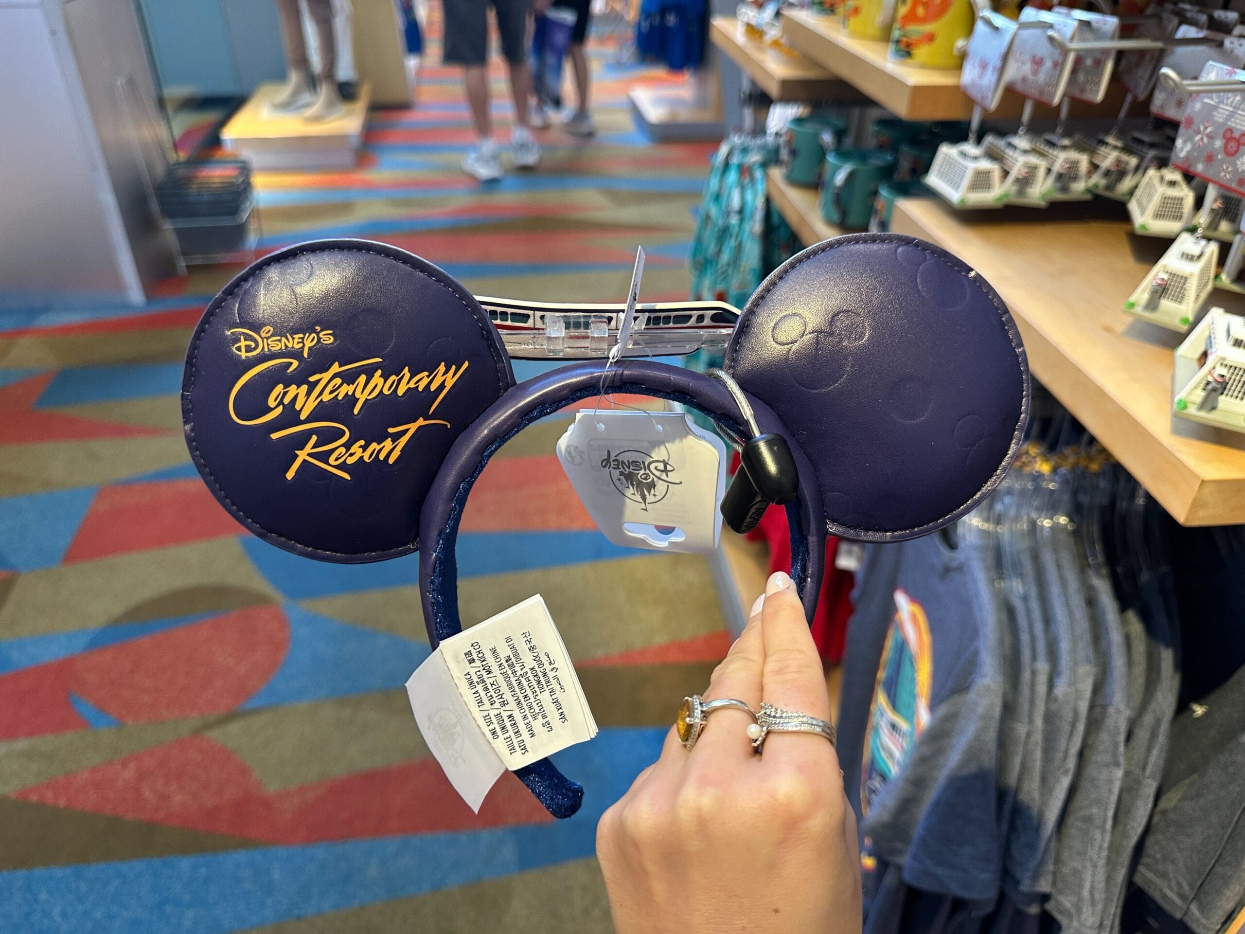 A hand displays Mickey ears labeled "Disney's Contemporary Resort" amid shirts and souvenirs, typical of a Walt Disney World shop.