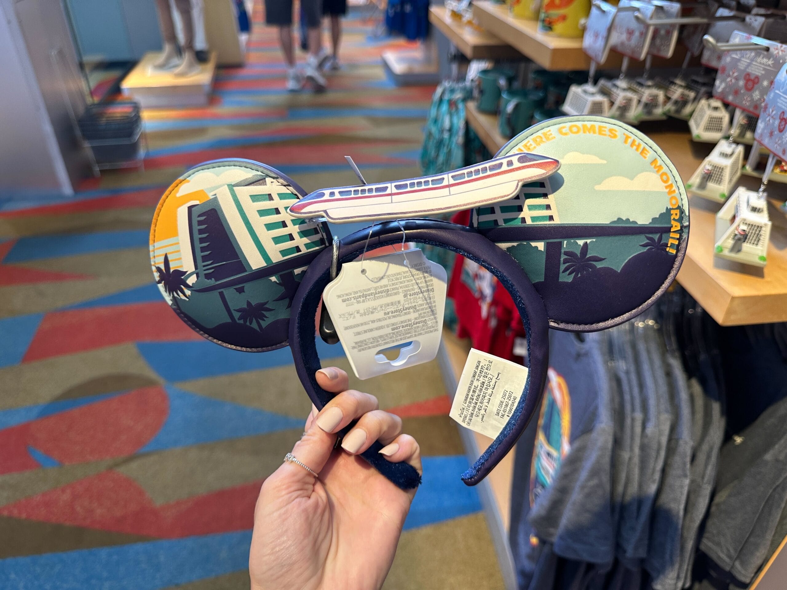 Hand holding a Moving Monorail Ear Headband in a vibrant Disney's Contemporary Resort shop, shirts and shelves in background.