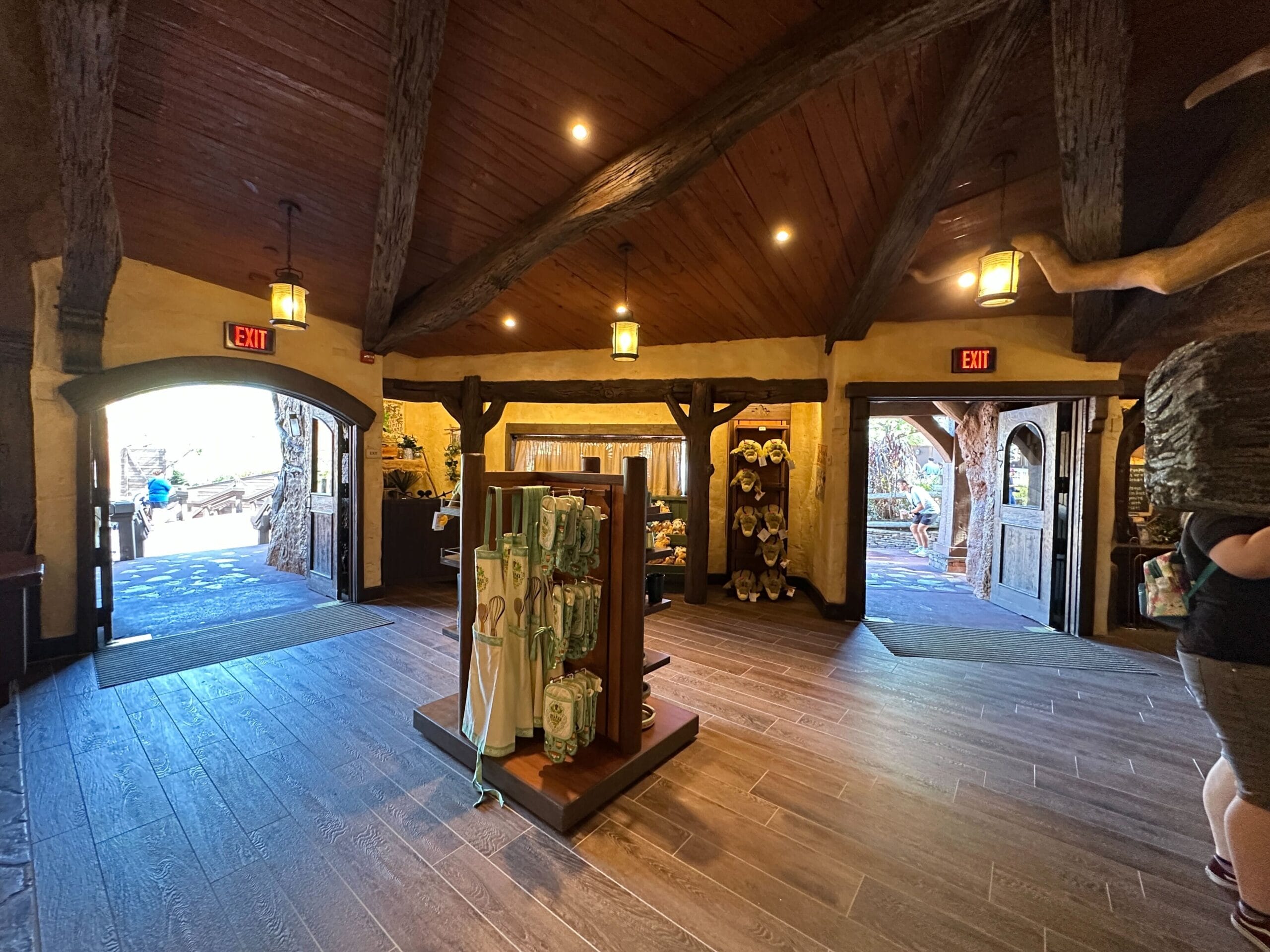 A cozy Magic Kingdom gift shop with wood floors, open doorways, hanging lights, and shelves of Disney merchandise—no castle in view.