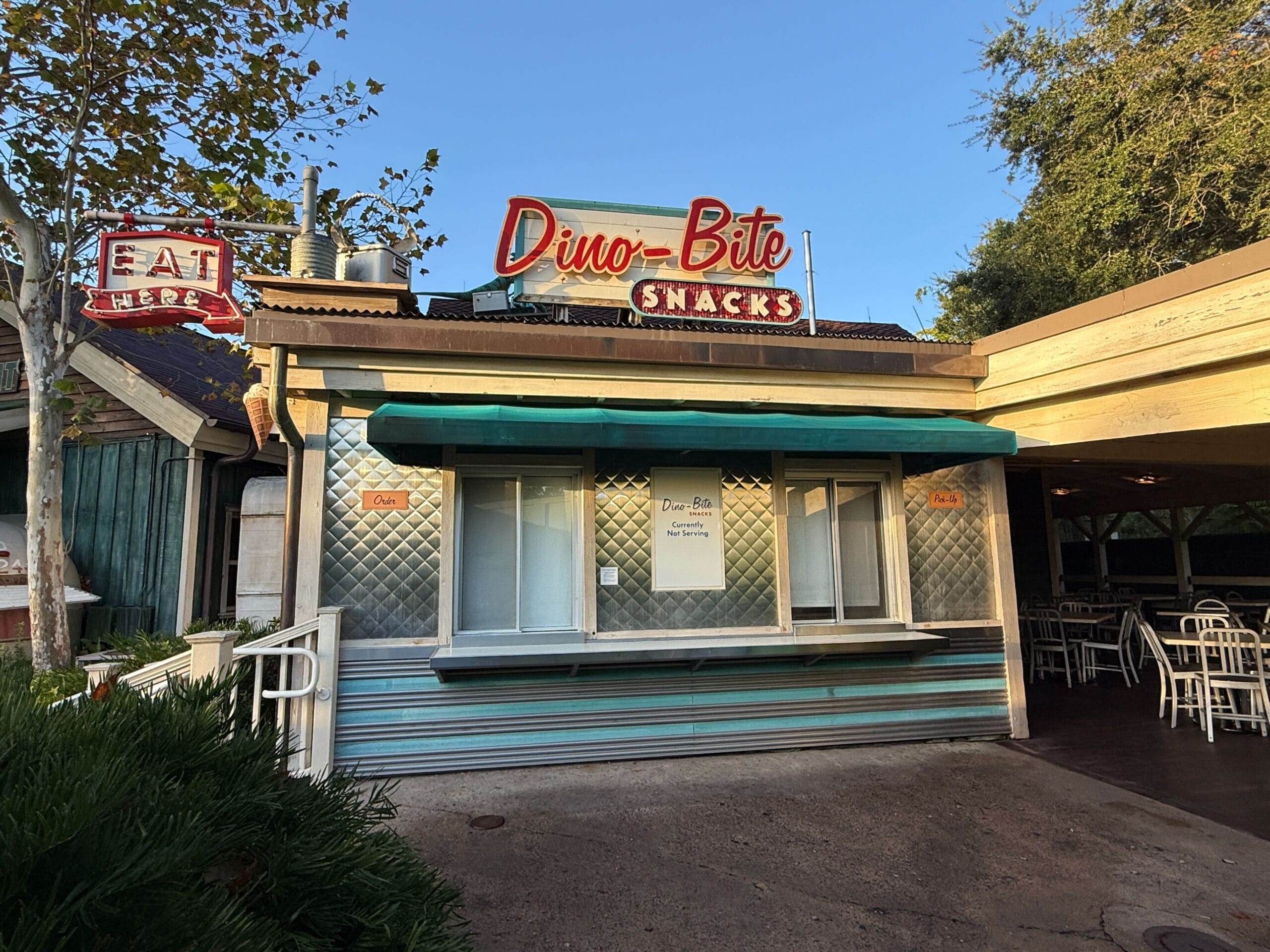A retro Dino-Bite Snacks stand at a Disney park, window closed, sign visible, nestled among trees and outdoor seating areas.