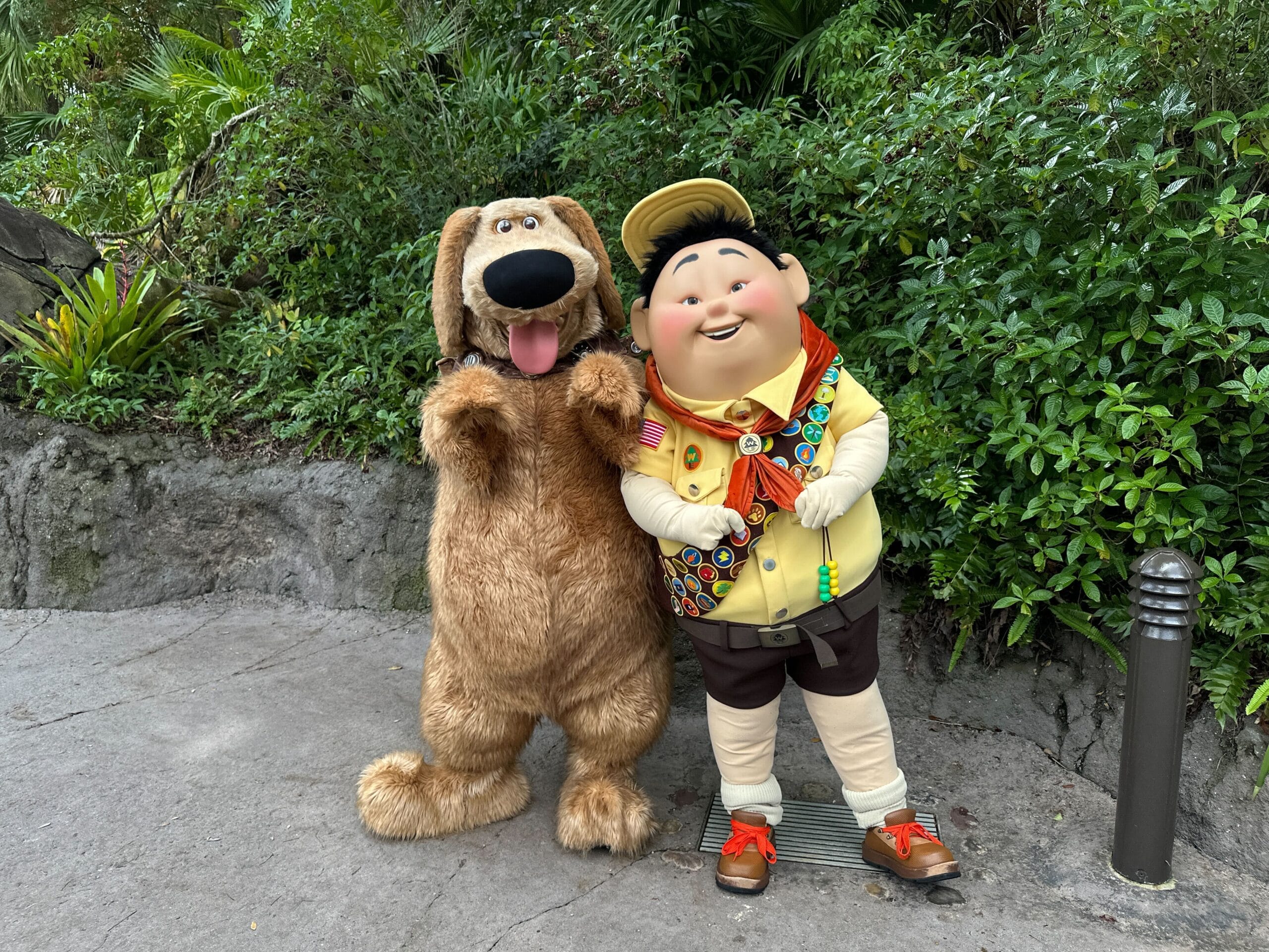 A character in a playful dog costume and a scout character stand together on a lush Disney park path filled with greenery.