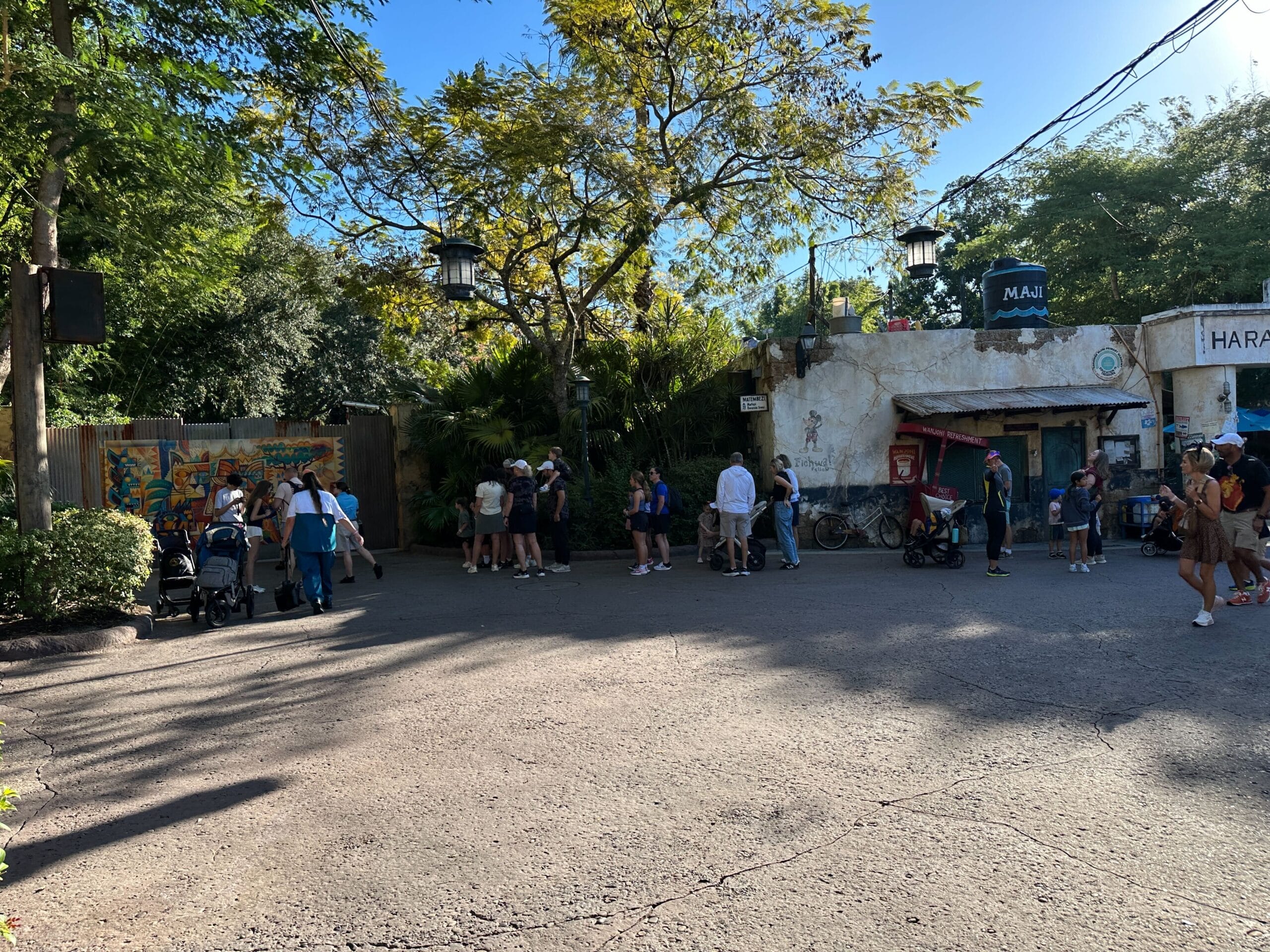 Guests line up outside a rustic building with trees, reminiscent of a cozy queue area in a Disney park, under sunny skies.
