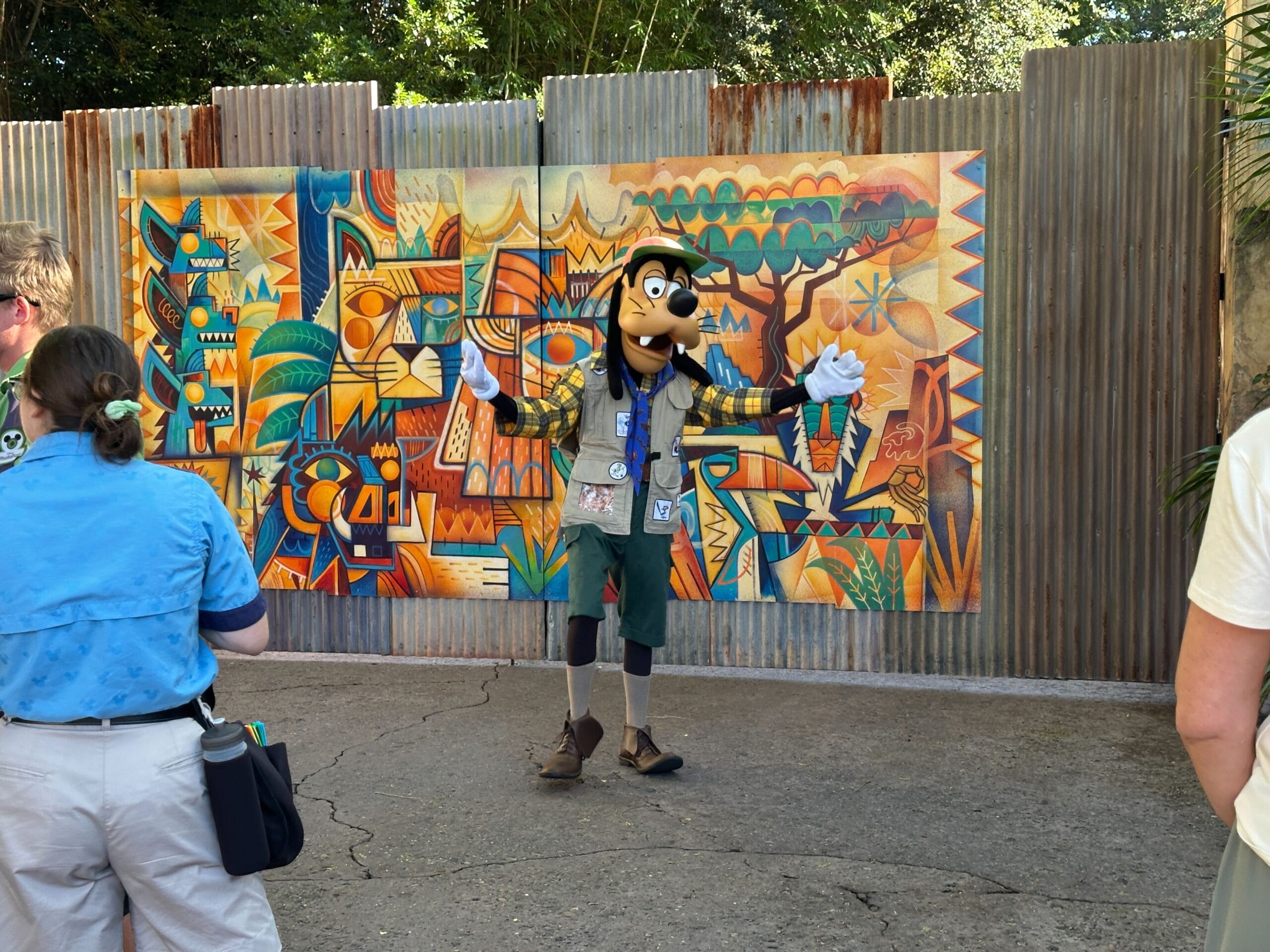 Goofy in safari gear poses for photos outside at a Disney park, with a vibrant jungle mural as the backdrop. No castle in view.